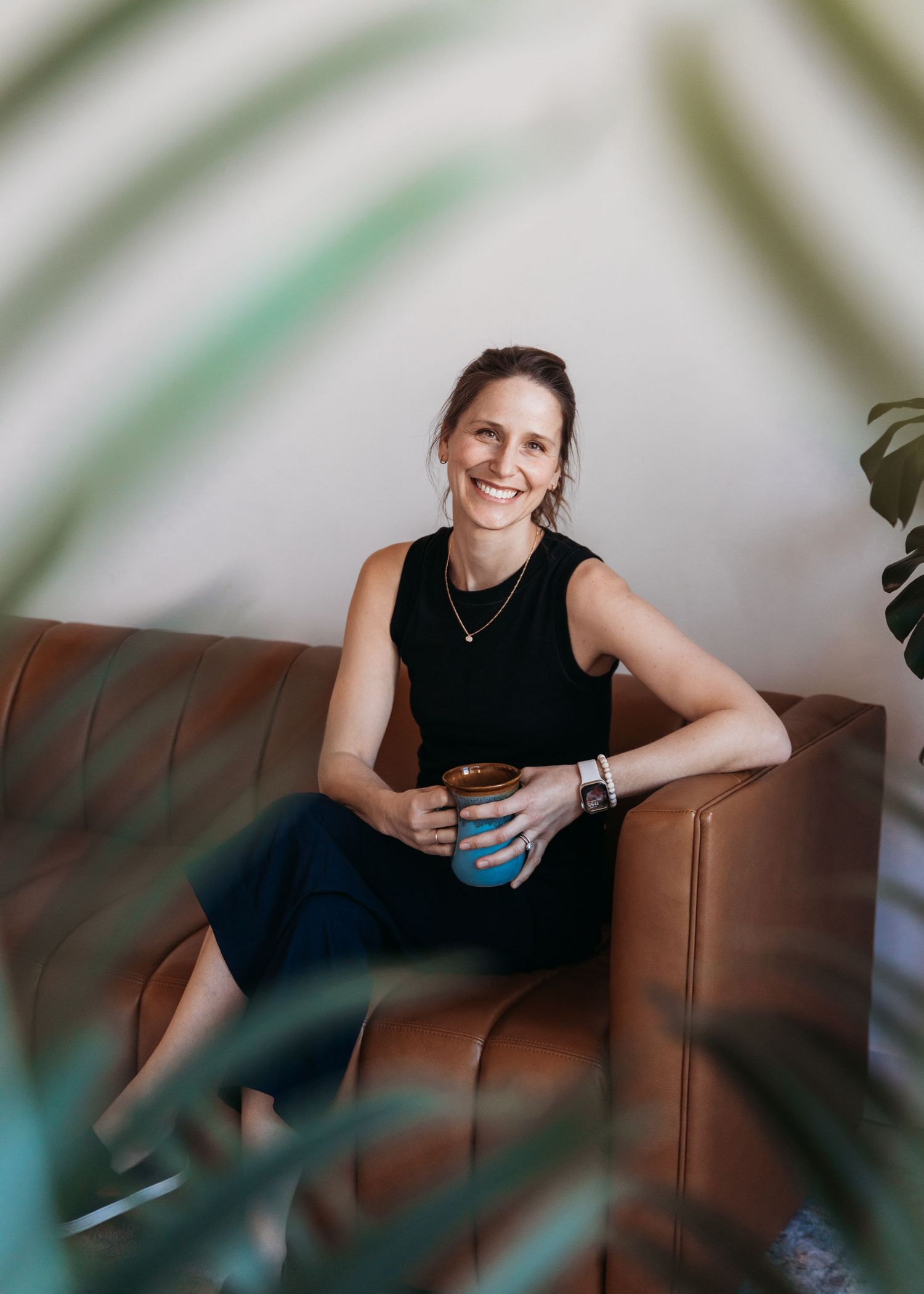 A person relaxes on a brown leather couch holding a blue coffee mug, with indoor plants visible in the foreground.