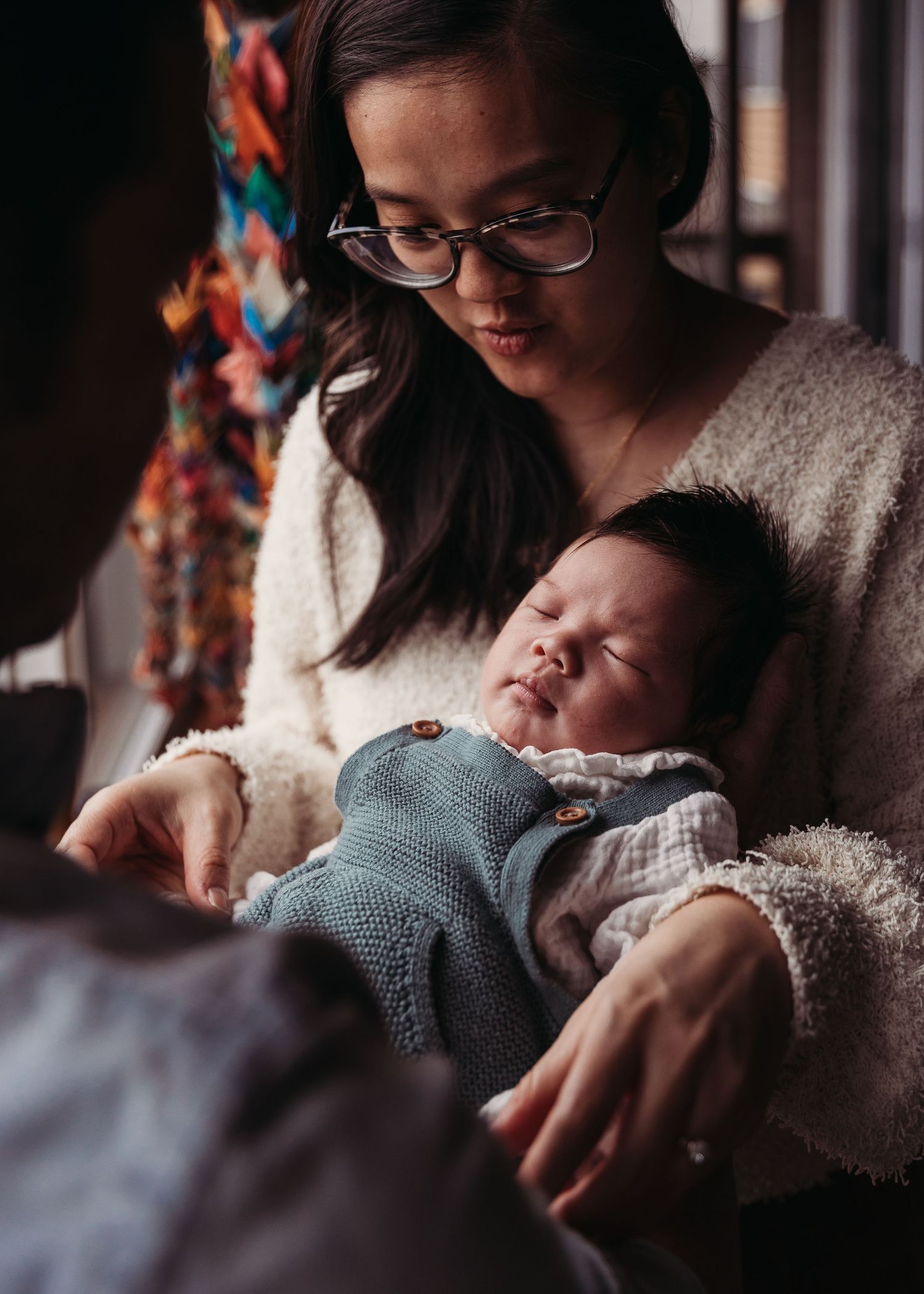 A parent in a cozy sweater cradles a sleeping baby wearing a soft blue knit blanket in warm lighting.