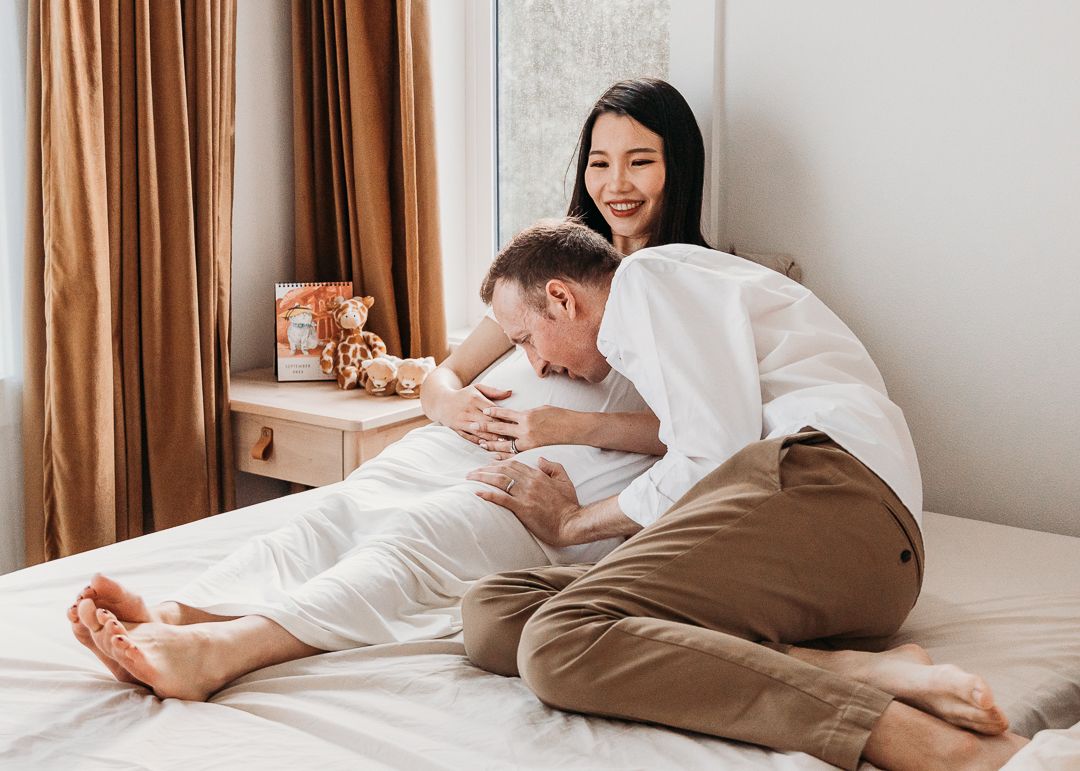 Happy couple in white and beige clothing sitting on bed together sharing a joyful moment at home.
