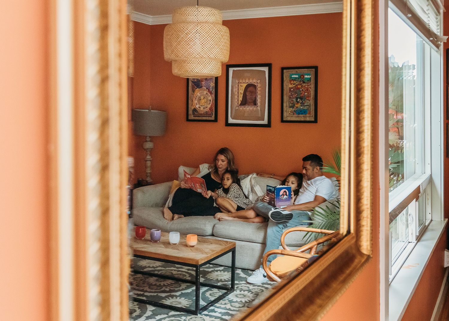 A cozy living room with orange walls, framed artwork, and people relaxing on a gray sofa near large windows.