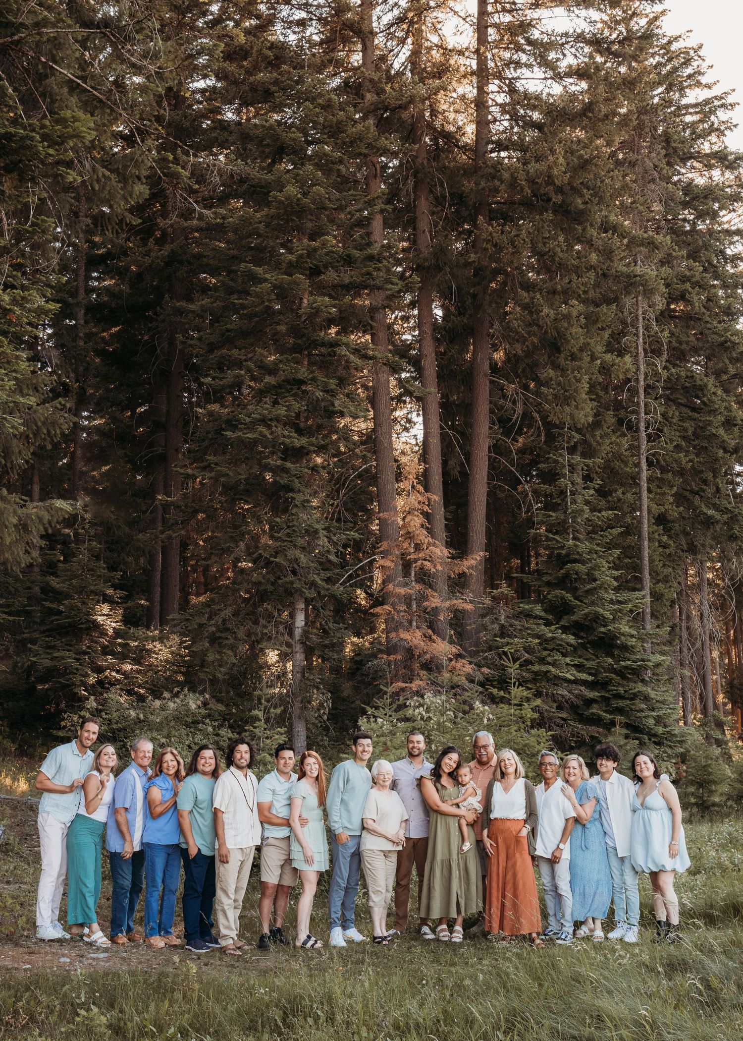 A large group poses together in coordinated blue and neutral outfits against a backdrop of tall evergreen trees.