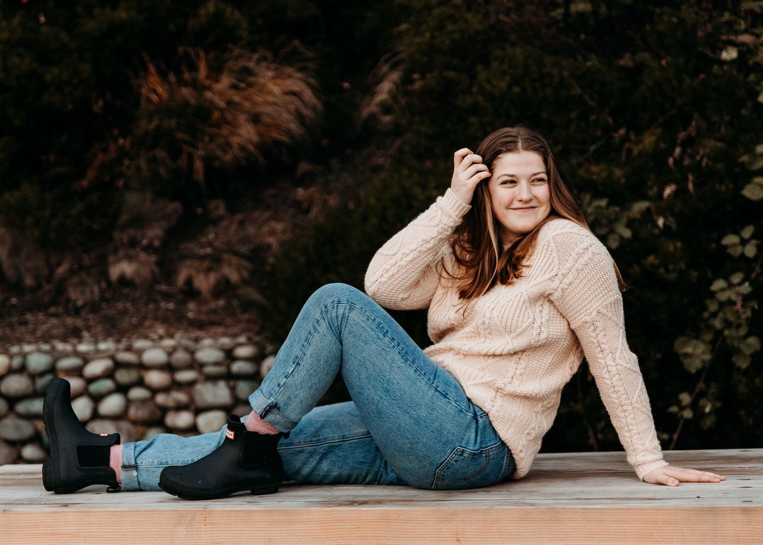 Person in a beige knit sweater and blue jeans sits casually on a stone retaining wall in an outdoor autumn setting.