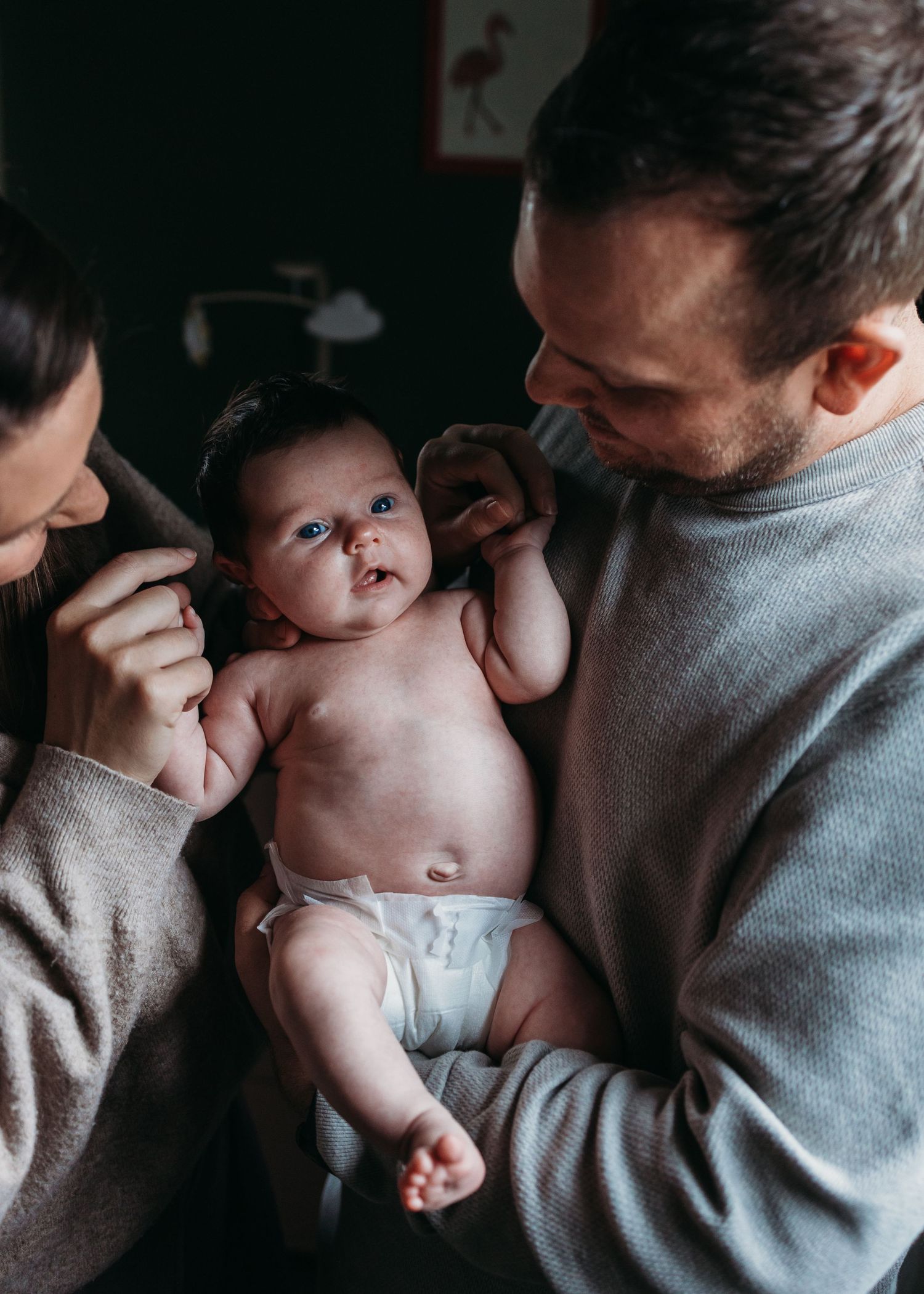 A tender moment shows parents gazing lovingly at their infant wearing a white diaper in moody lighting.