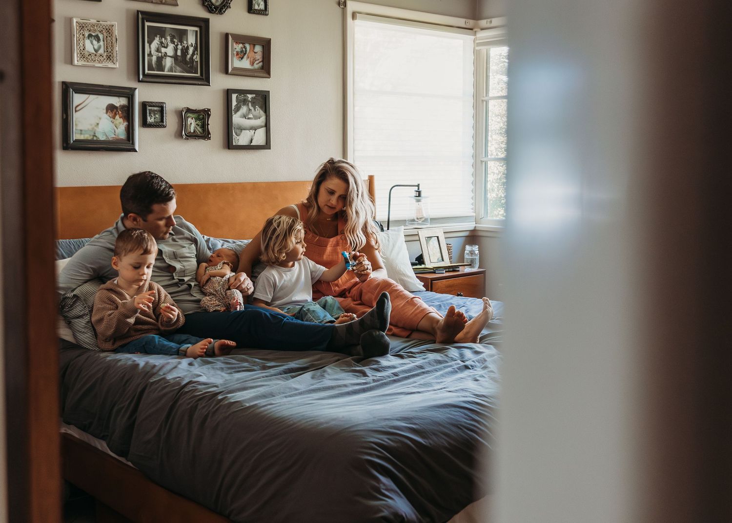 A family relaxes together on a bed in a cozy bedroom with natural light and framed photos on the wall.