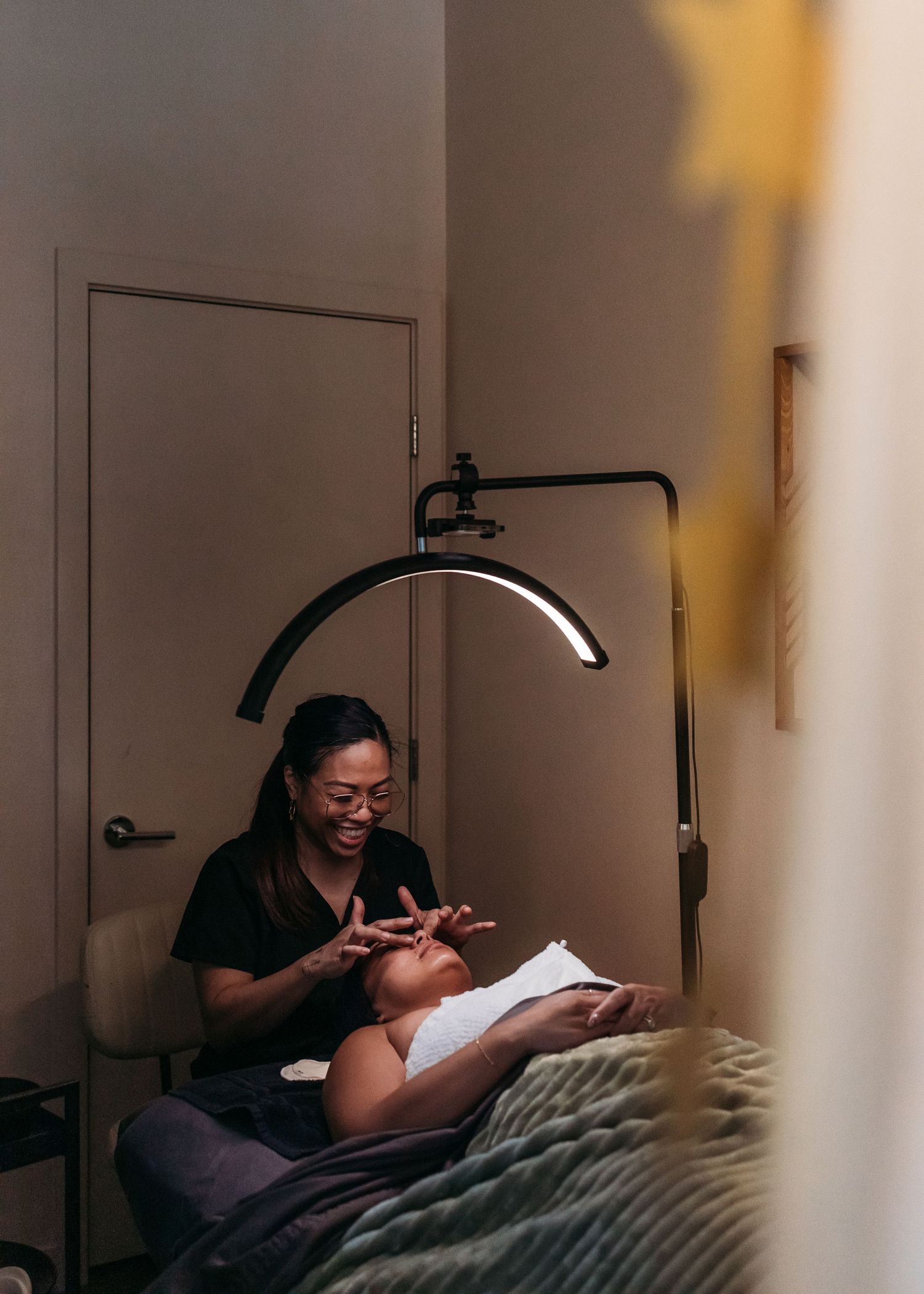 A person performs a facial treatment on a client under a curved task lamp in a dimly lit spa room.