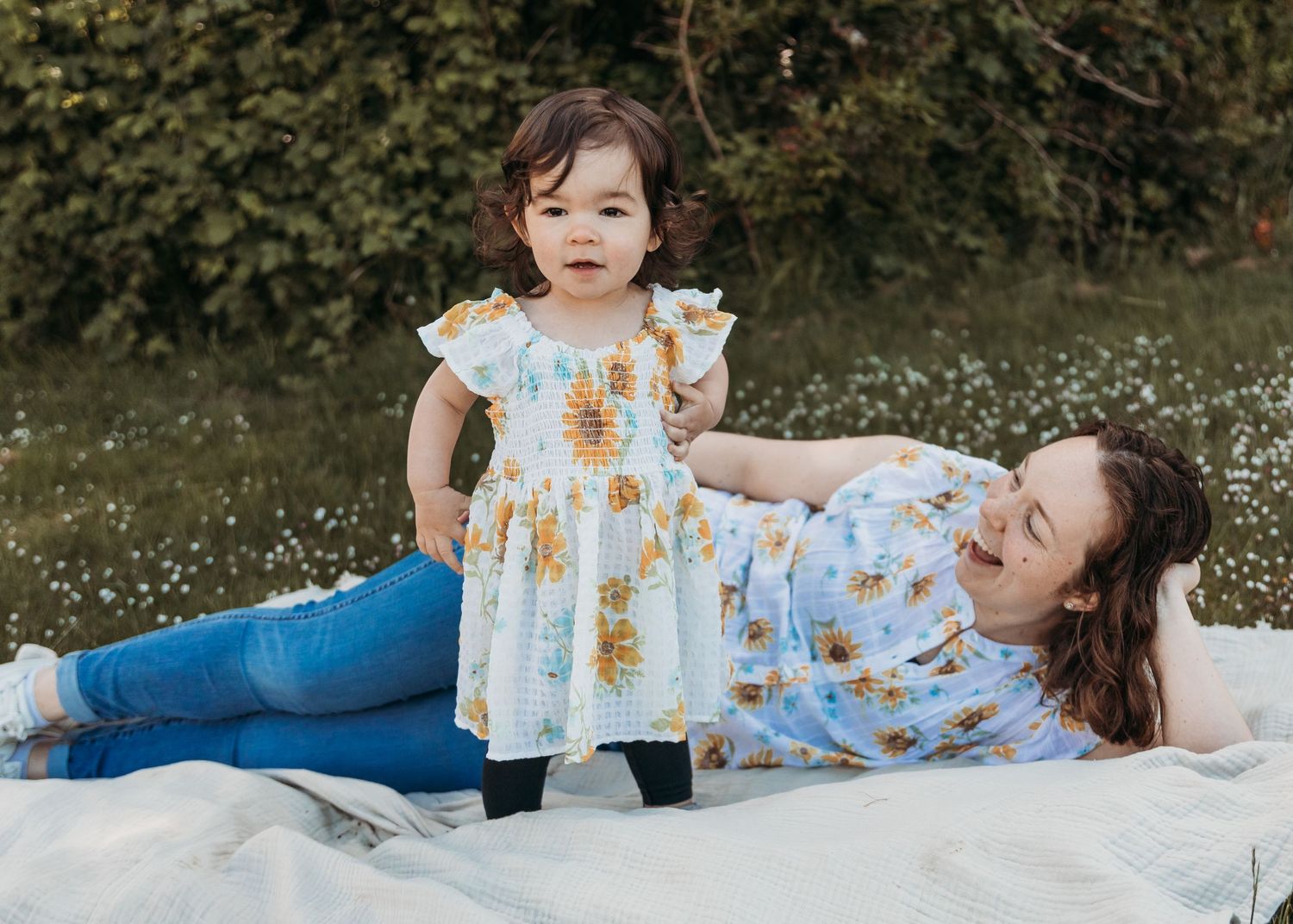 Happy parent and toddler laying on white blanket outdoors wearing matching blue floral dresses in garden setting.