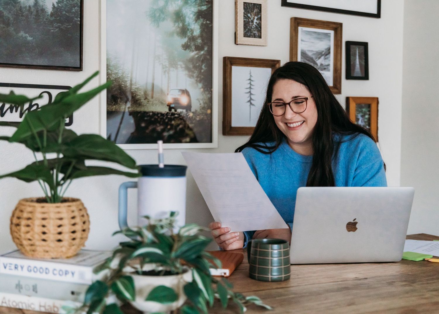 Remote worker in comfortable home office setup with laptops, houseplants and framed artwork on wall.