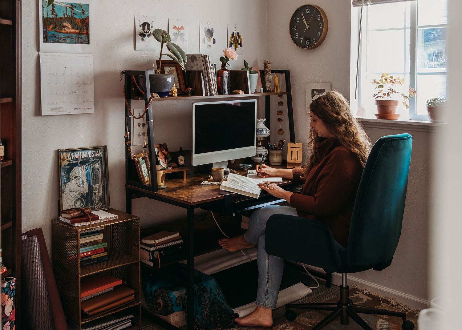 A person sits at a wooden computer desk with bookshelves and decorative items in a cozy home office setup.