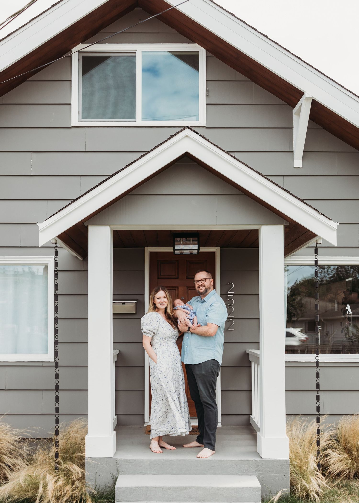 Couple standing in the covered entryway of a modern gray house with ornamental grasses in the landscaping.
