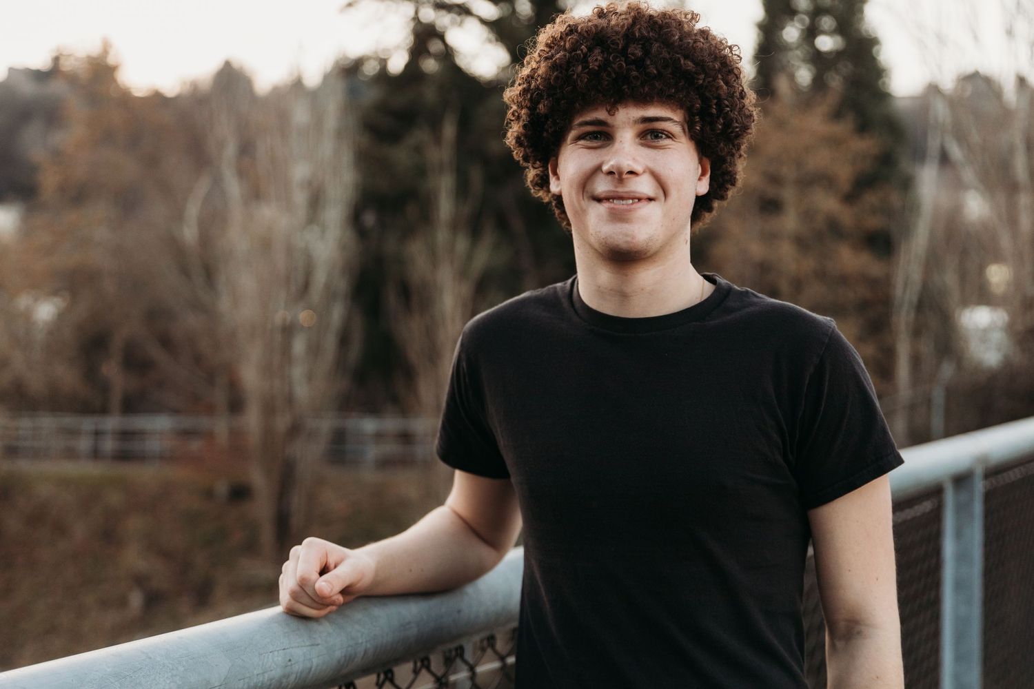 Someone wearing a black t-shirt and curly hair smiles while leaning against a railing outdoors with trees in background.
