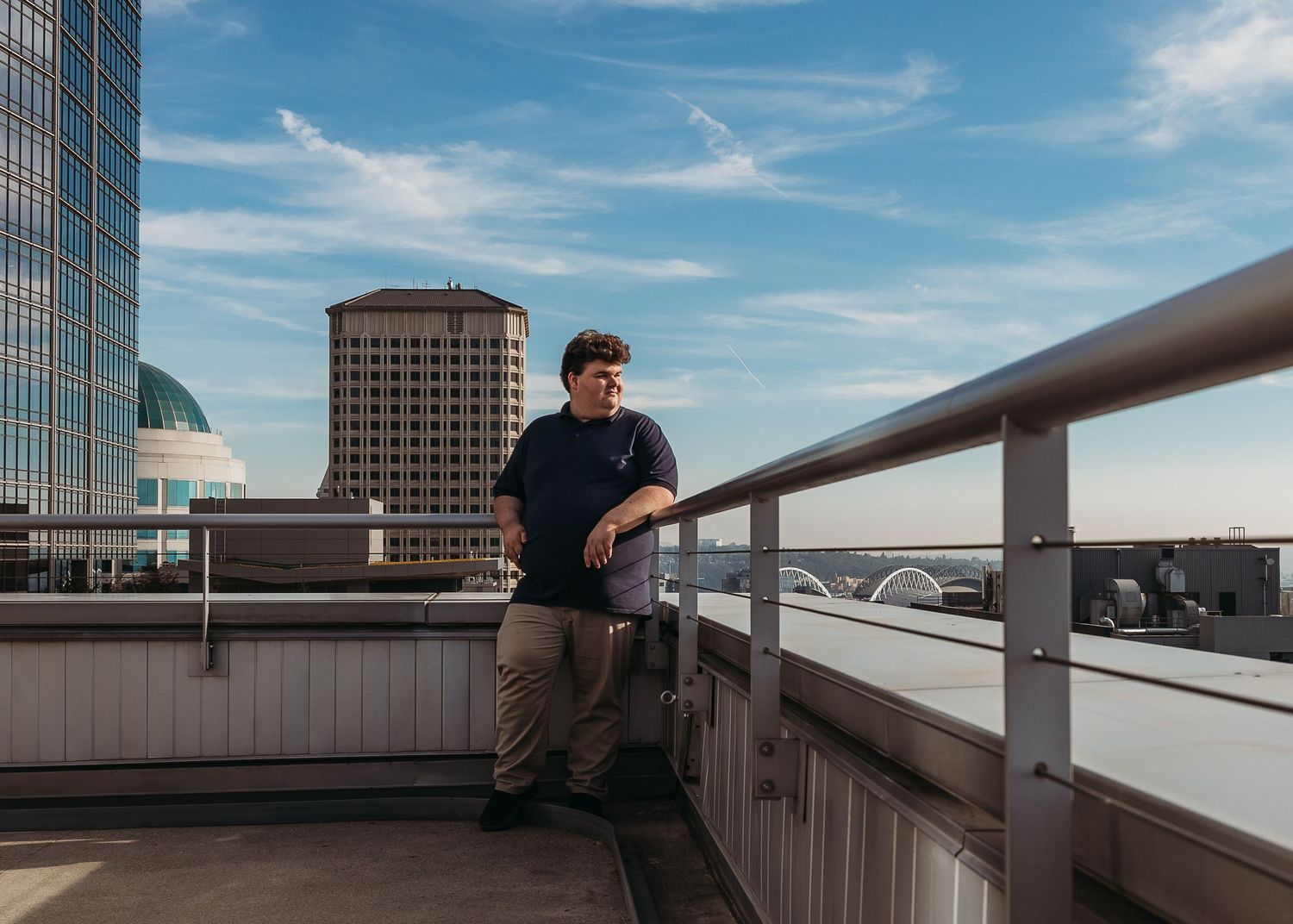 Person stands on an urban rooftop terrace with metal railings and city buildings against a blue sky in the background.