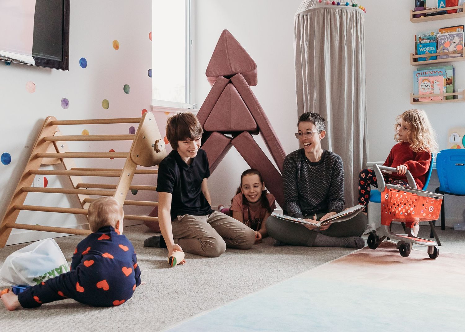 Parents and children relax together in a playroom with wooden climbing equipment and soft furnishings in muted tones.