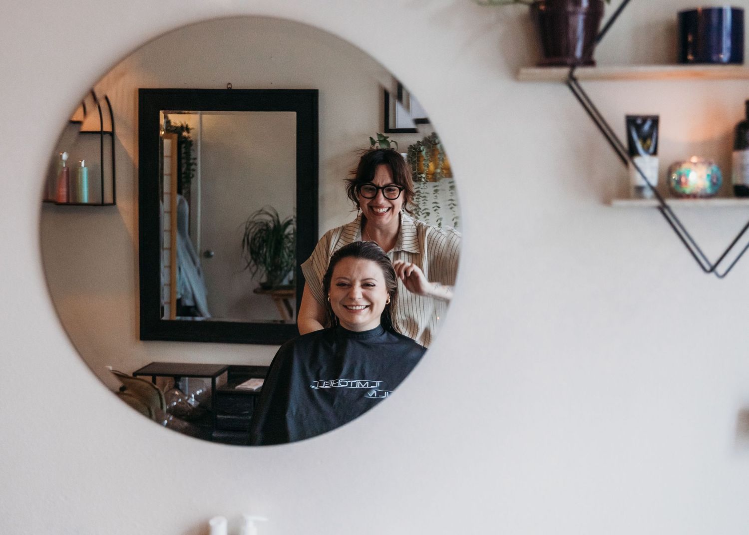 Circular mirror reflection shows a haircut in progress at a cozy home salon with rustic floating shelves on the wall.