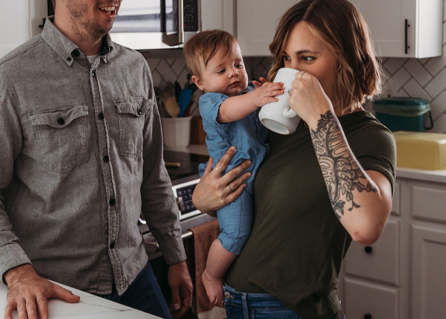 A casual family moment in the kitchen with a child between two people while one takes a drink from a cup.