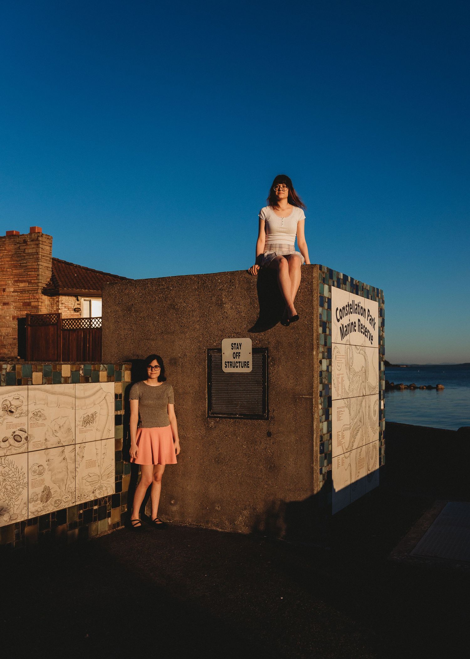 Two people dressed in summer attire pose against a concrete wall at sunset near the ocean.
