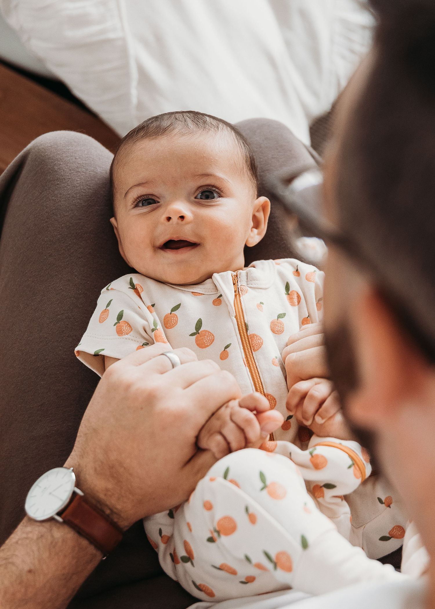 Smiling baby wearing white and orange patterned onesie being held by adult hands with wristwatch.