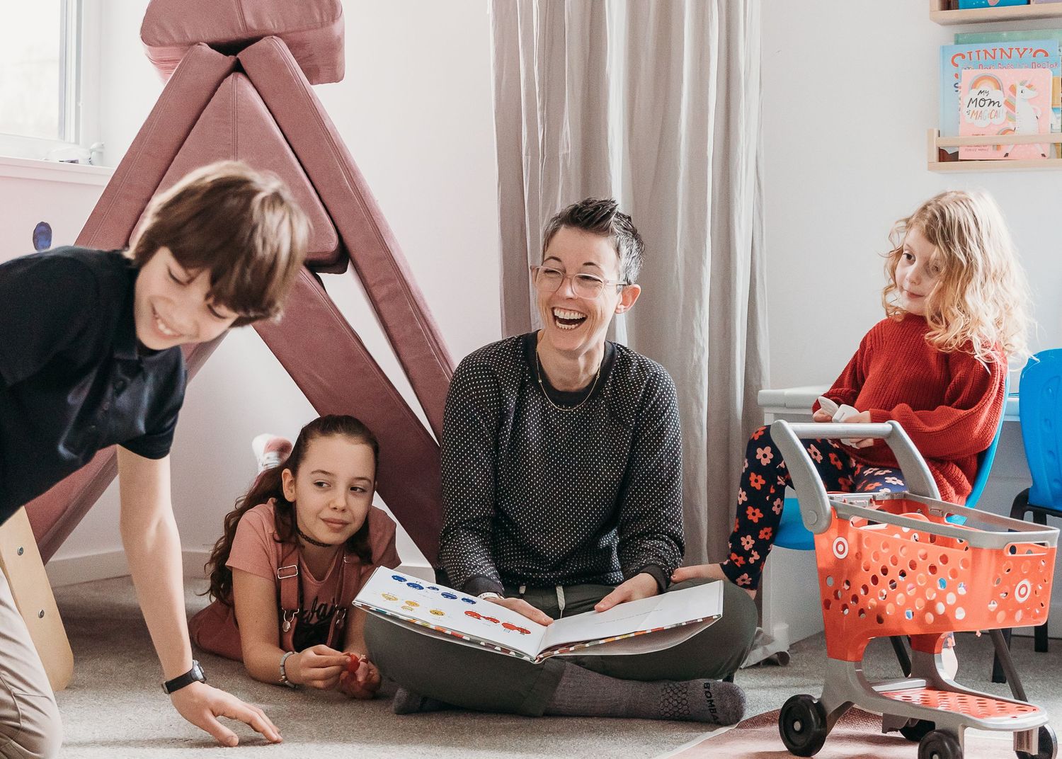 A group sits together reading a book and laughing while enjoying quality time in a playroom with a pink tent.