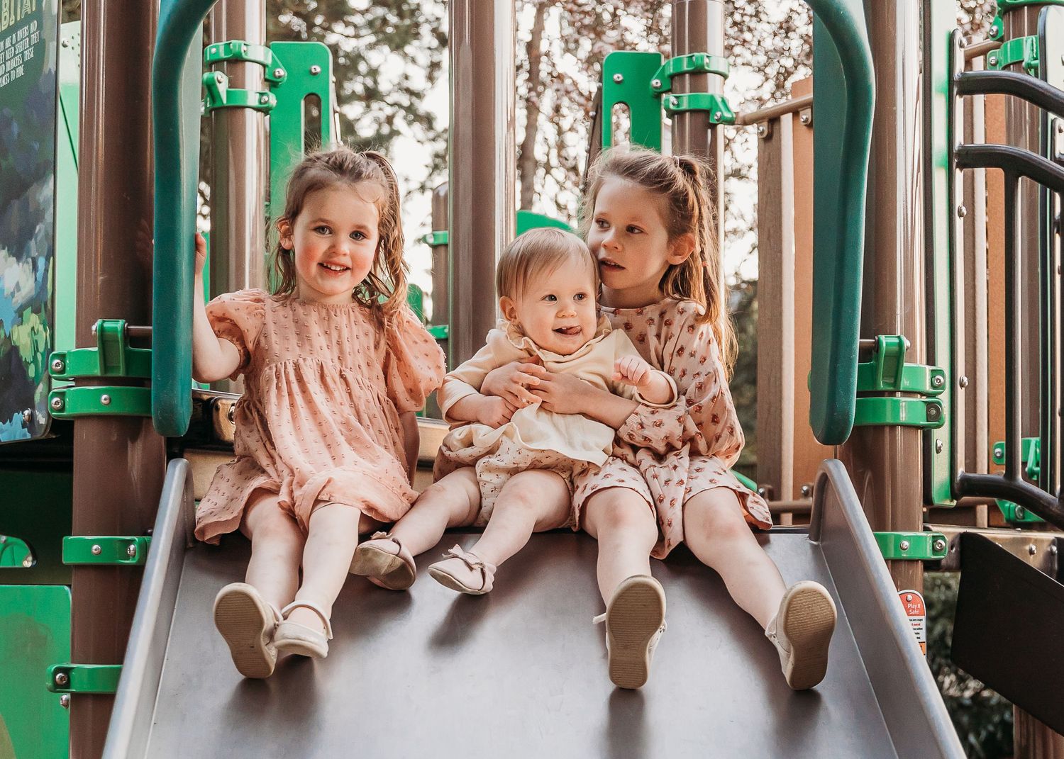 Three children sit together on a playground slide with green metal equipment on a sunny day.