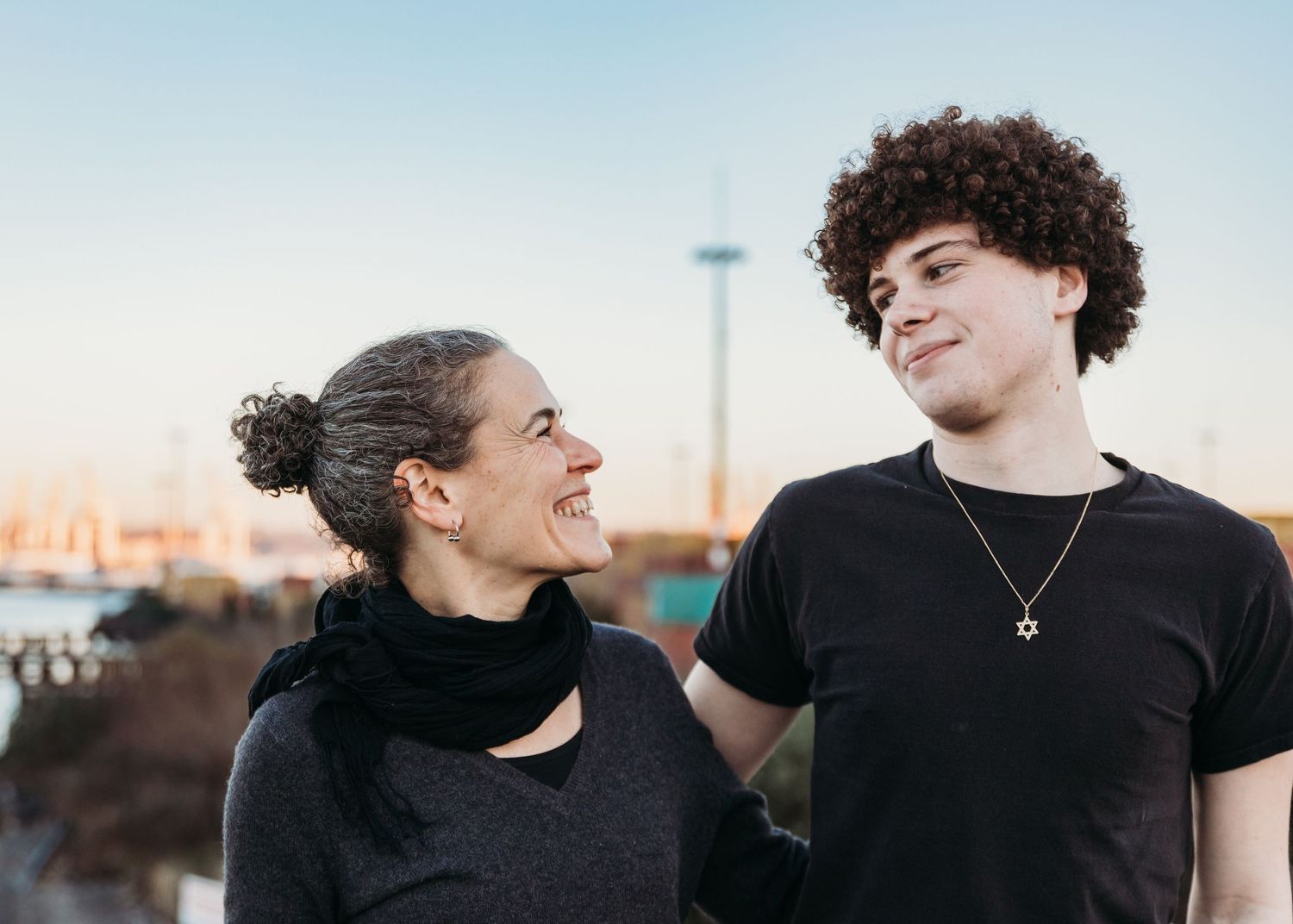 A young couple share a playful moment together in casual attire against an urban sunset backdrop.