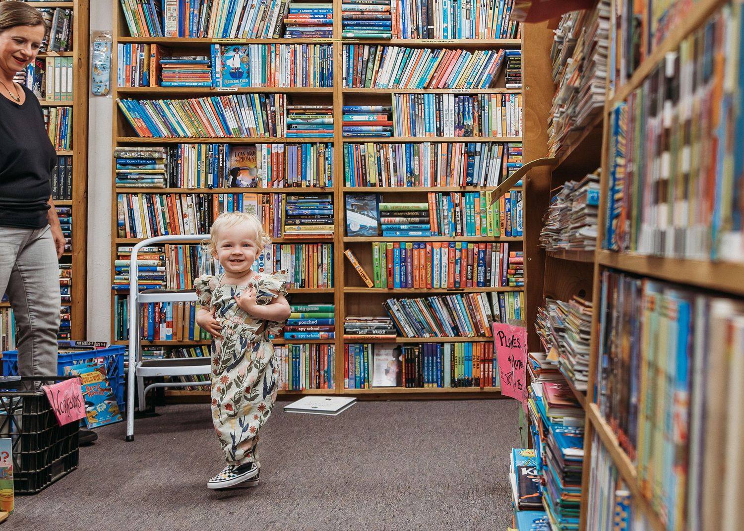 A toddler explores a cozy bookstore aisle surrounded by tall bookshelves filled with colorful books.
