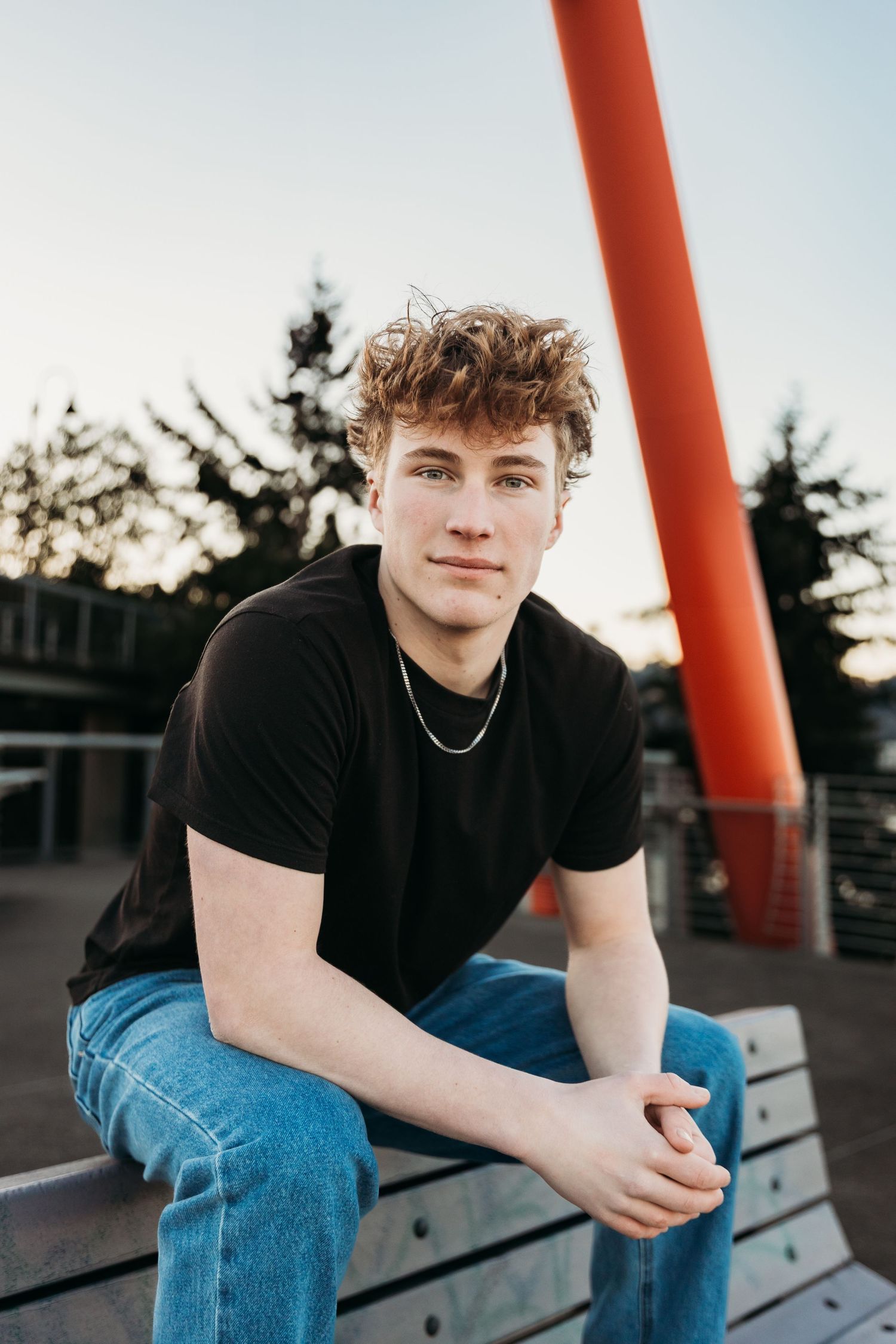 Young individual in black t-shirt and jeans sits pensively on bench near large red architectural pillar at sunset.