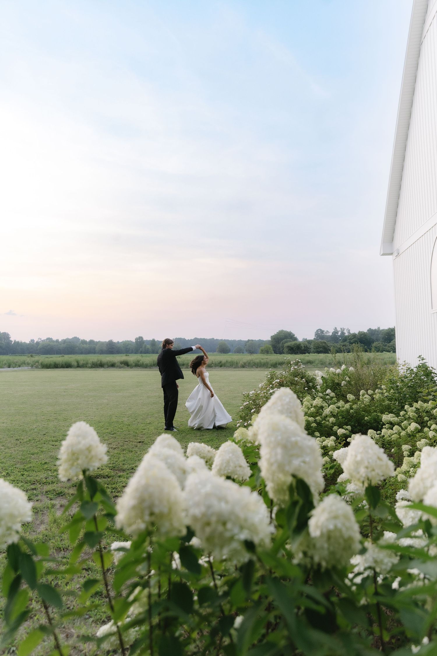Bride and groom dancing at sunset in a green field, with white hydrangeas in the foreground near a white barn.