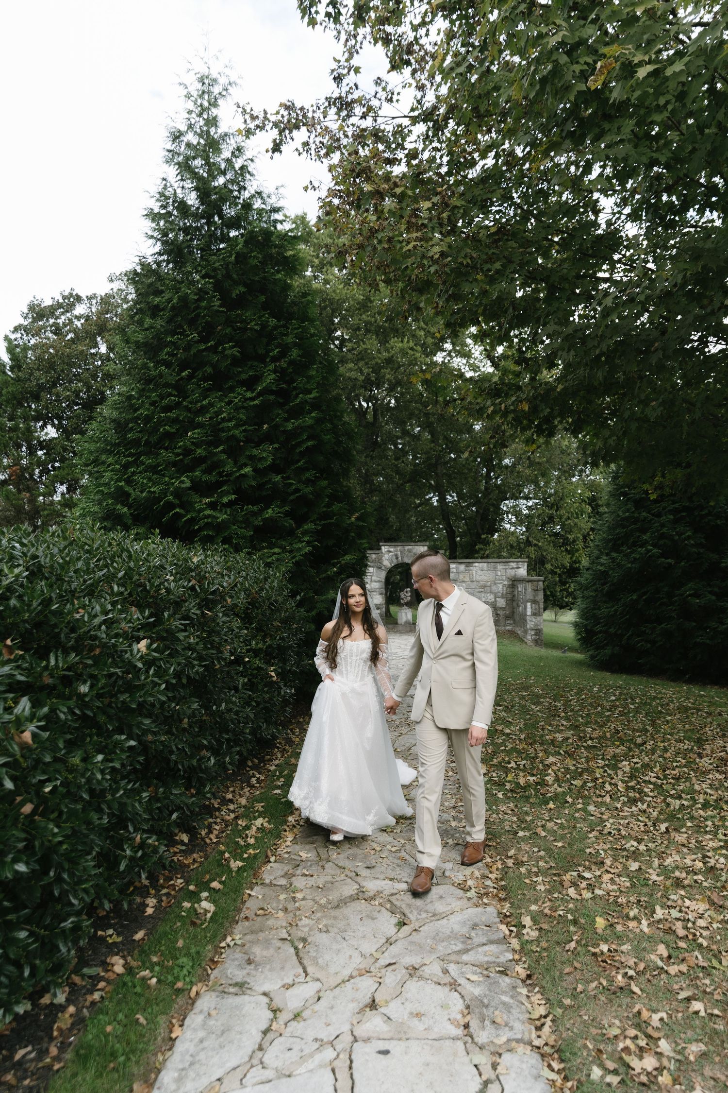A couple walks hand in hand along a stone path surrounded by autumn leaves and evergreen trees in a garden setting.