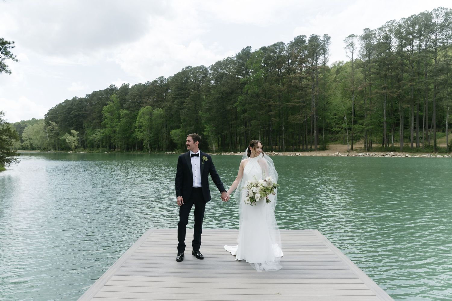 Bride and groom hold hands walking on a dock by a serene lake surrounded by lush green trees.