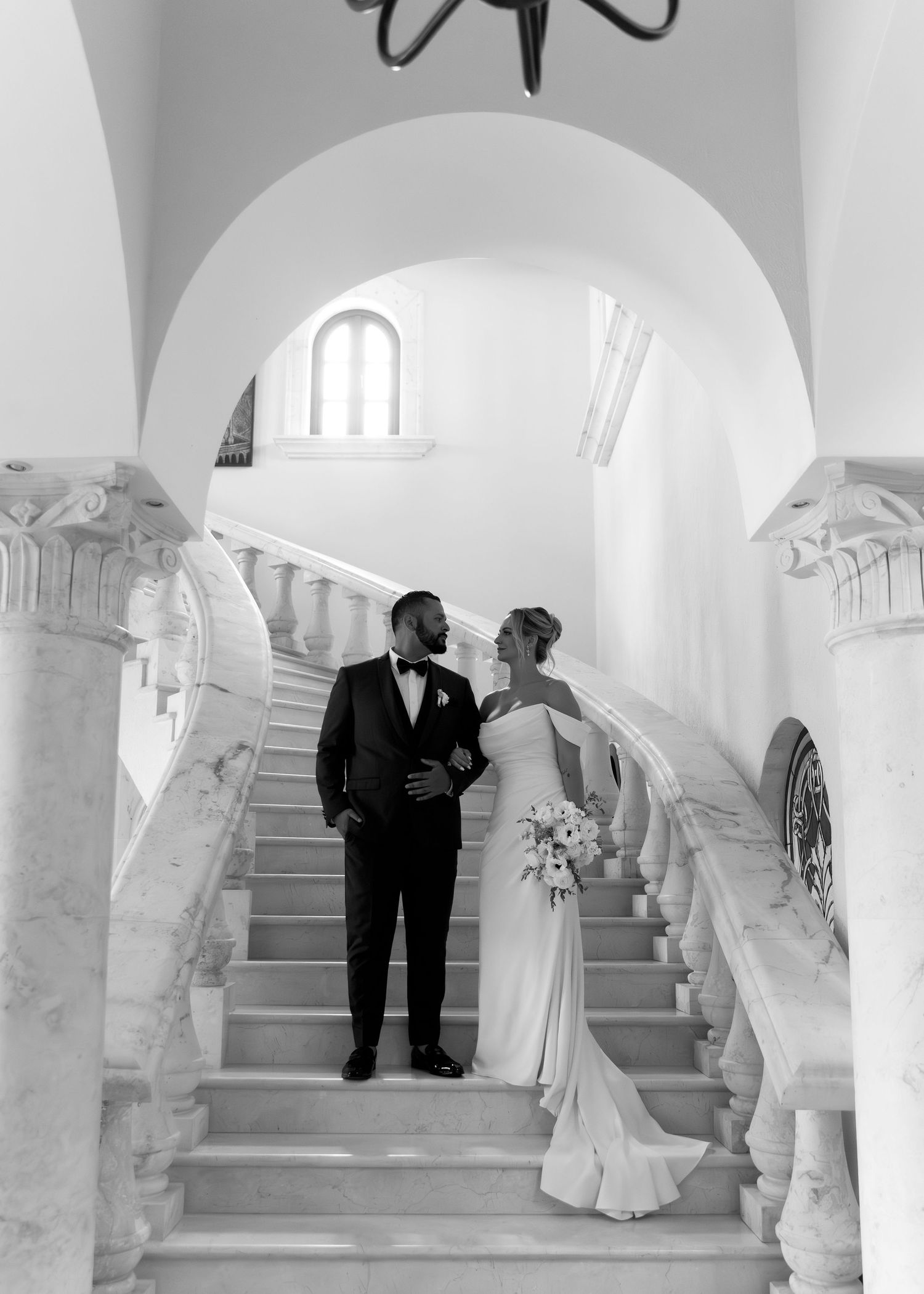 Black and white artistic wedding photograph showing couple in elegant attire on grand marble staircase with arches.