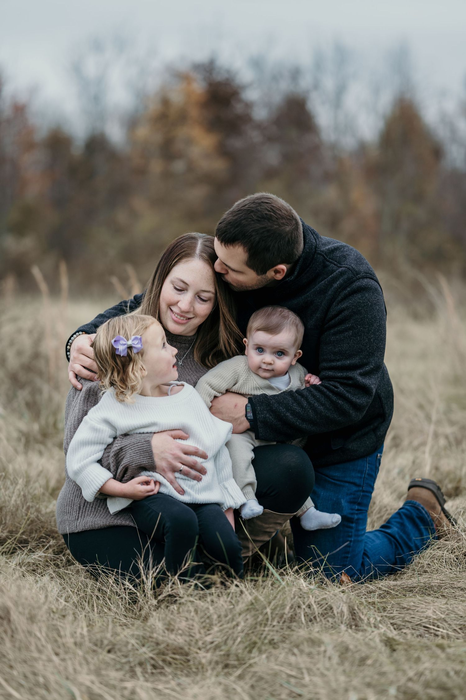 Family of four sitting in autumn field, parents cuddling toddler girl and baby outdoors.