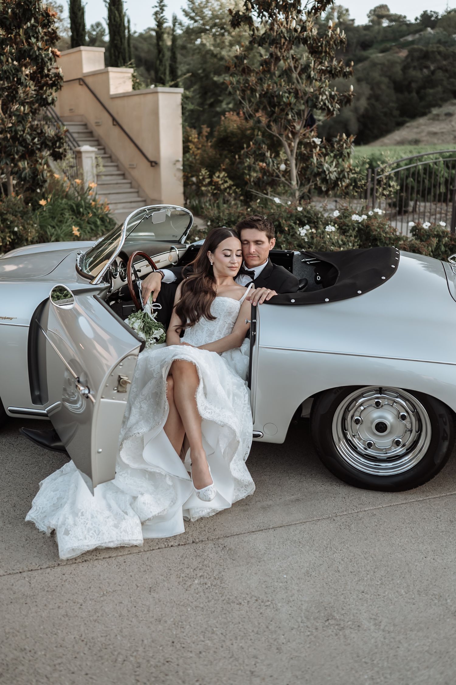 Bride in white gown sits in vintage silver convertible with groom, surrounded by lush greenery at outdoor wedding venue.