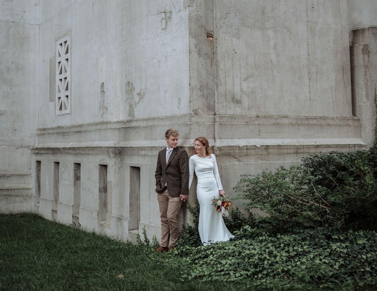 Couple in wedding attire embraces against white stone church wall with ivy growing in foreground.