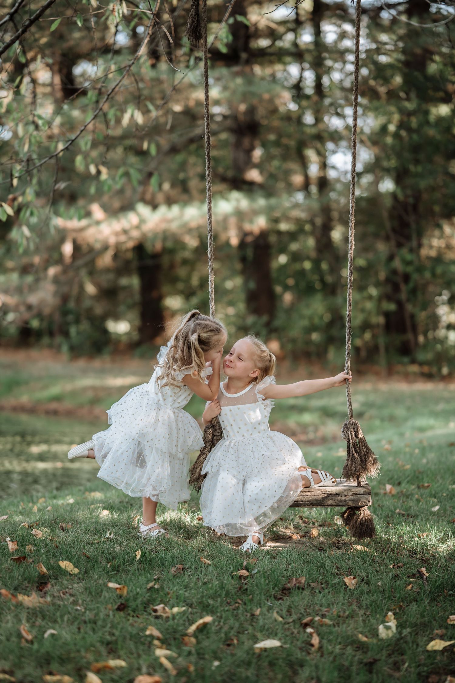 Two young girls in white dresses share a sweet moment on a rustic rope swing in a lush green garden.
