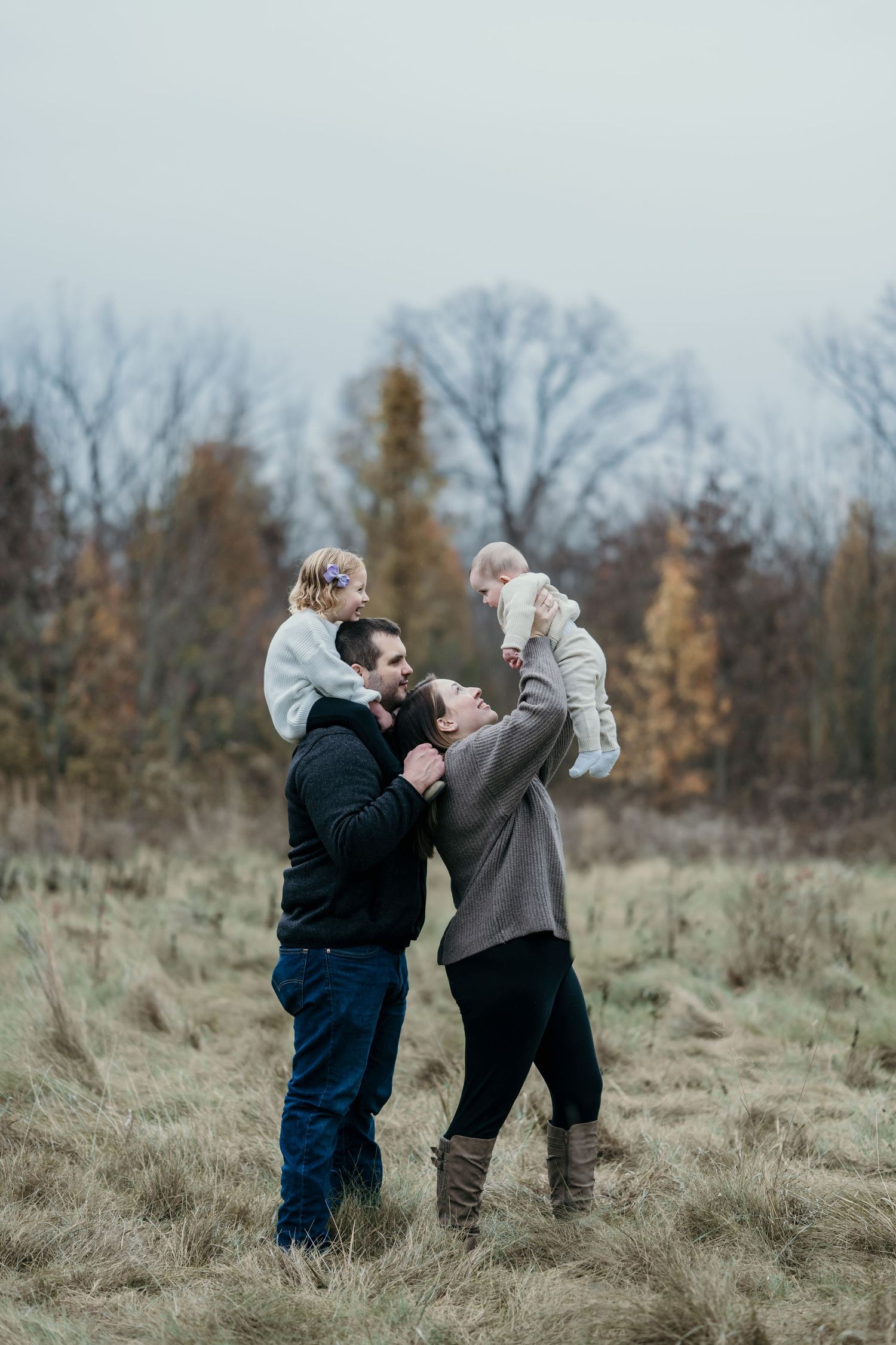 A family of four enjoys a playful autumn moment in a field, with parents lifting two young children outdoors.