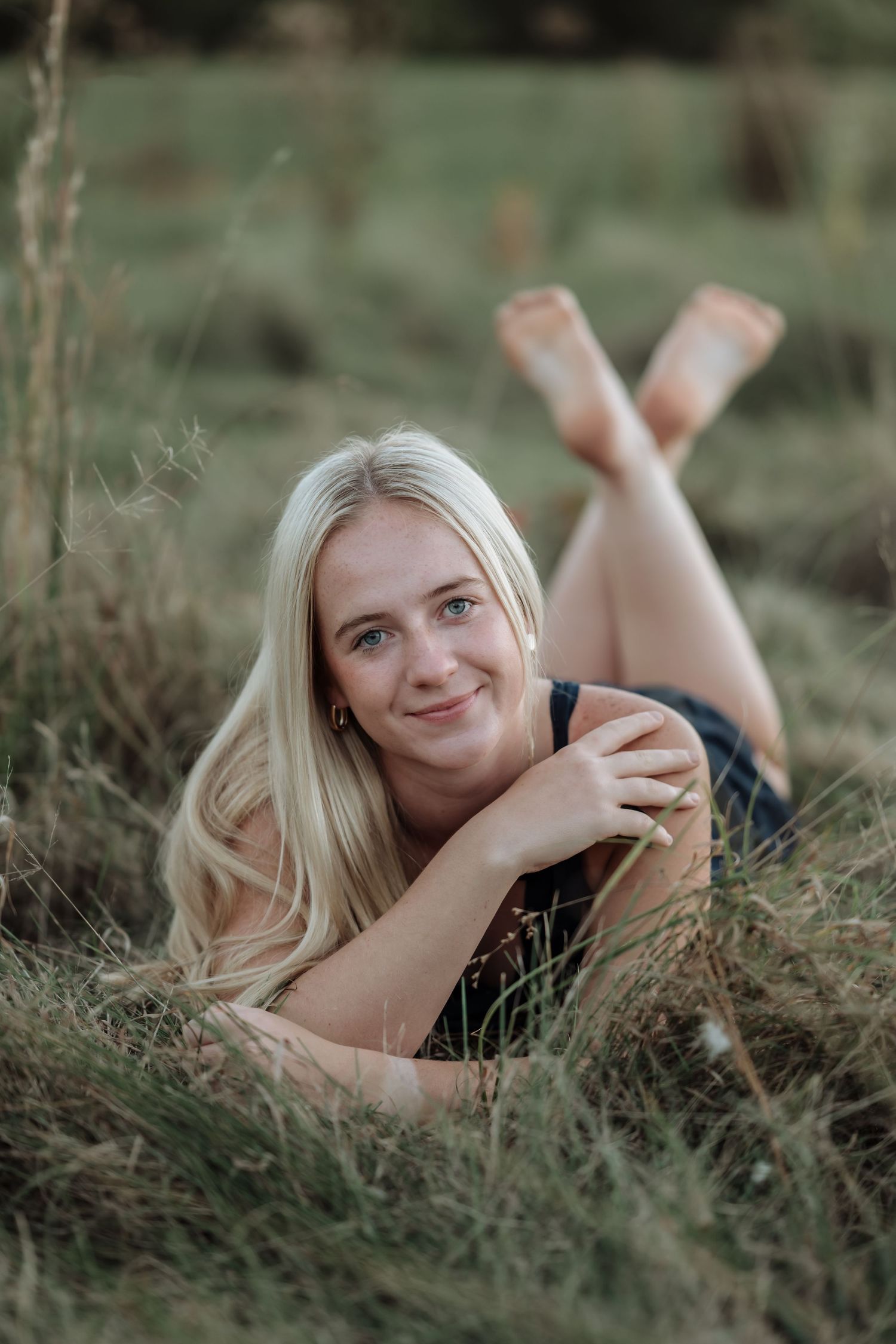 Smiling blonde woman lying in grass field, propped on elbows with crossed legs, enjoying a relaxed outdoor portrait.
