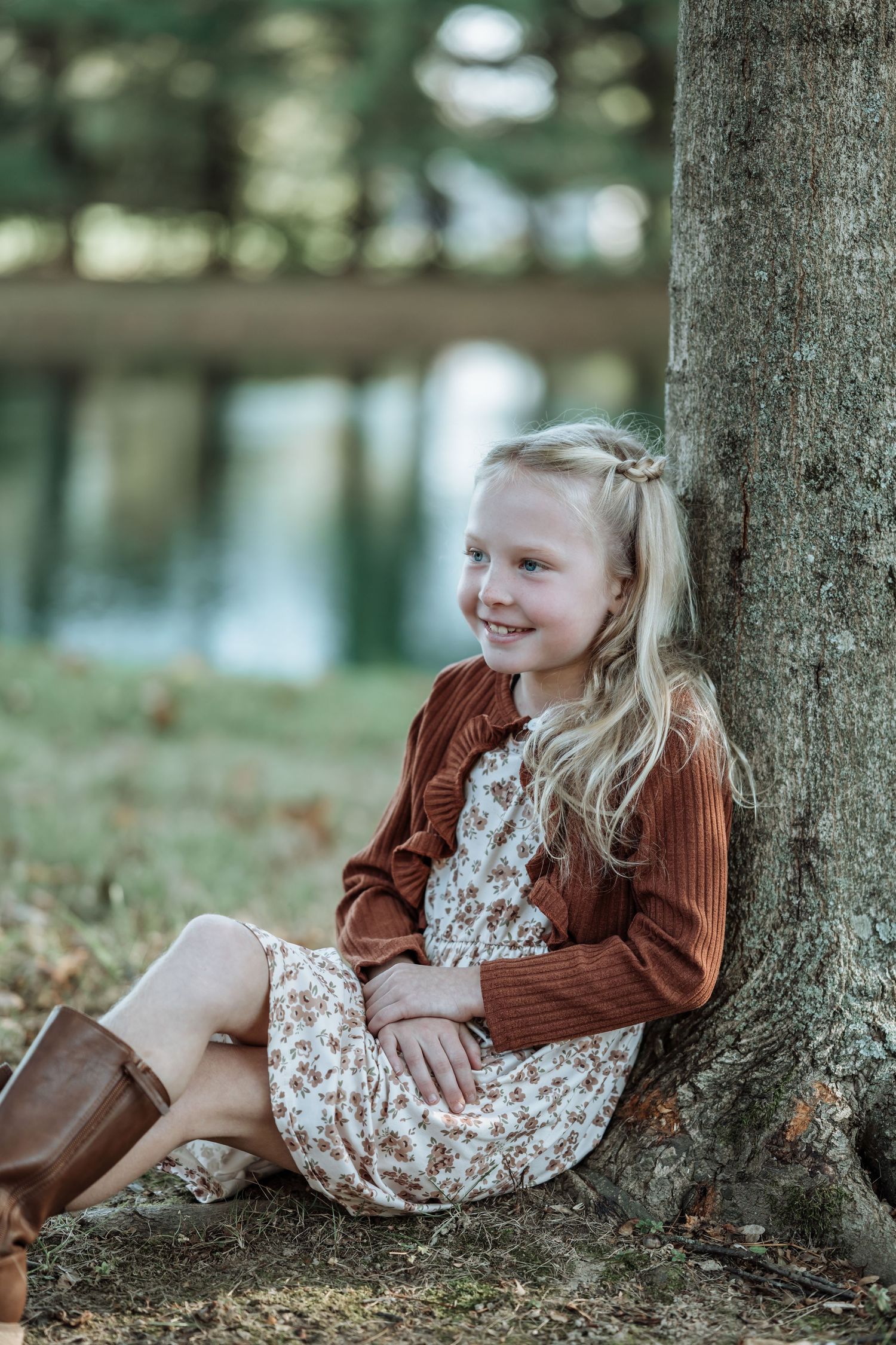 A child in a brown sweater and floral dress lies playfully against a tree trunk in an autumn forest setting.