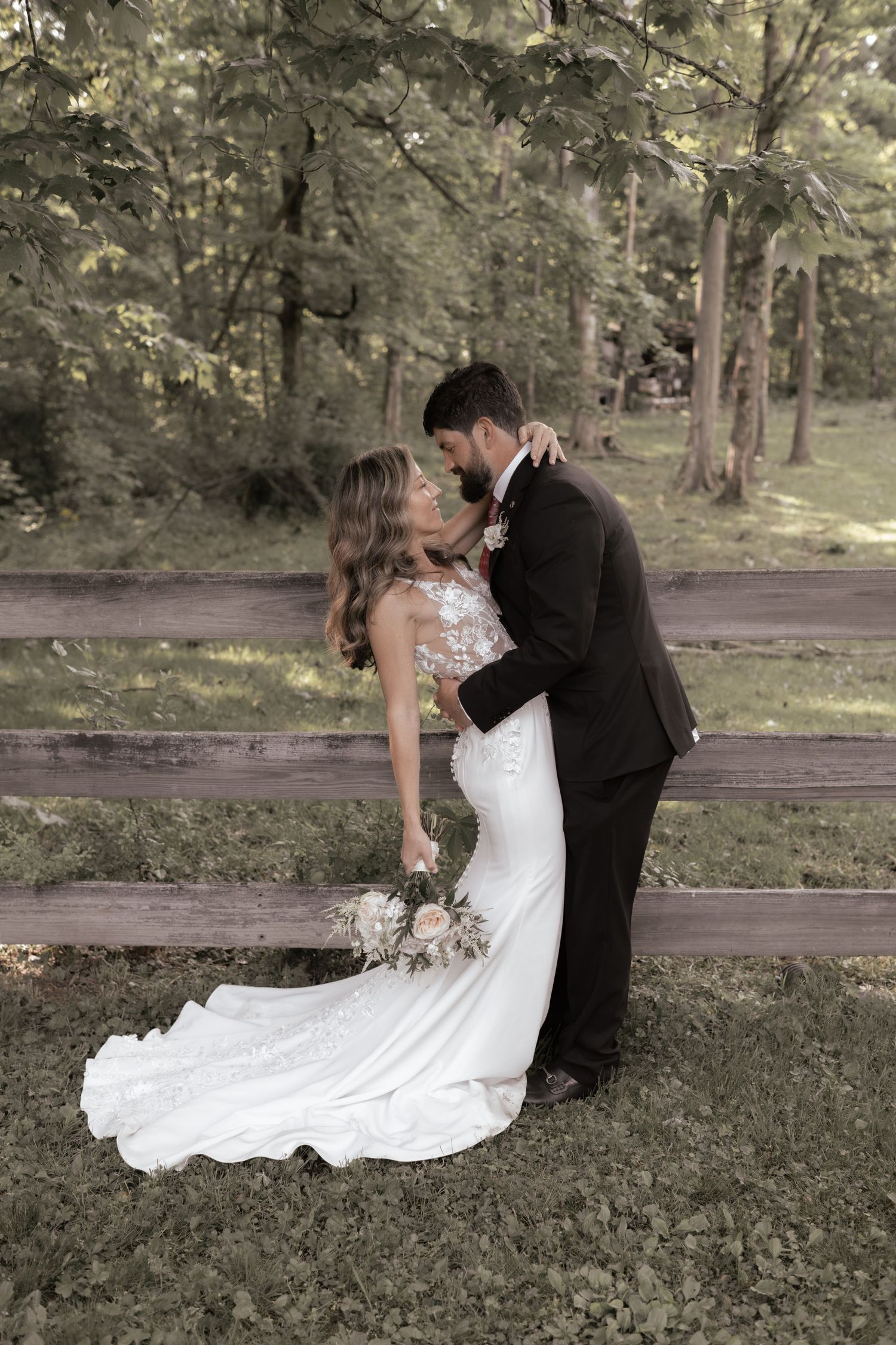 Bride and groom share an intimate moment along a rustic wooden fence in a dreamy woodland setting.