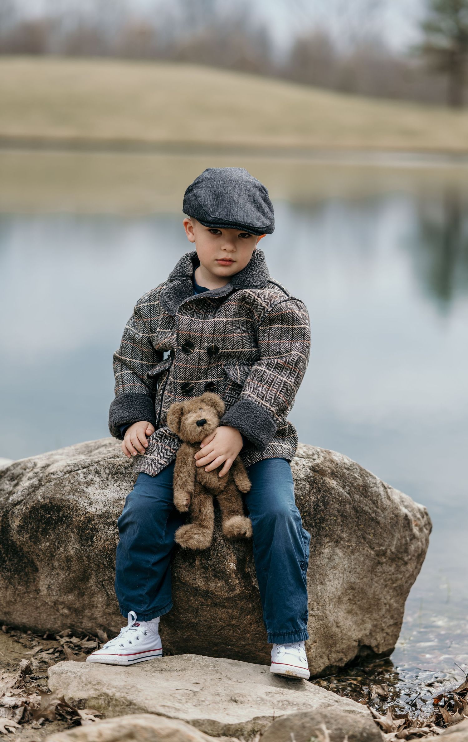 A young boy in a plaid coat and newsboy cap sits on a rock by a lake, holding a teddy bear.