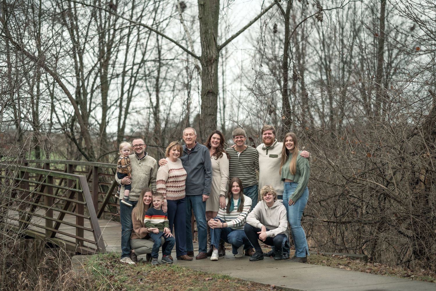 Large family group portrait outdoors on a path surrounded by bare winter trees, smiling together for a photo.