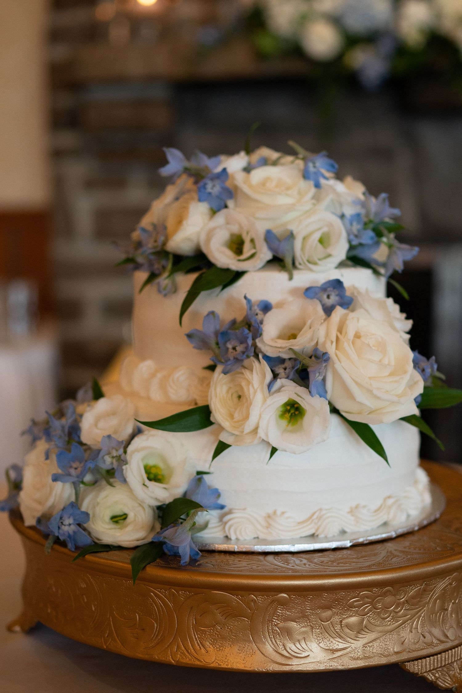 Wedding cake adorned with fresh white roses and delicate blue flowers on an ornate gold stand.