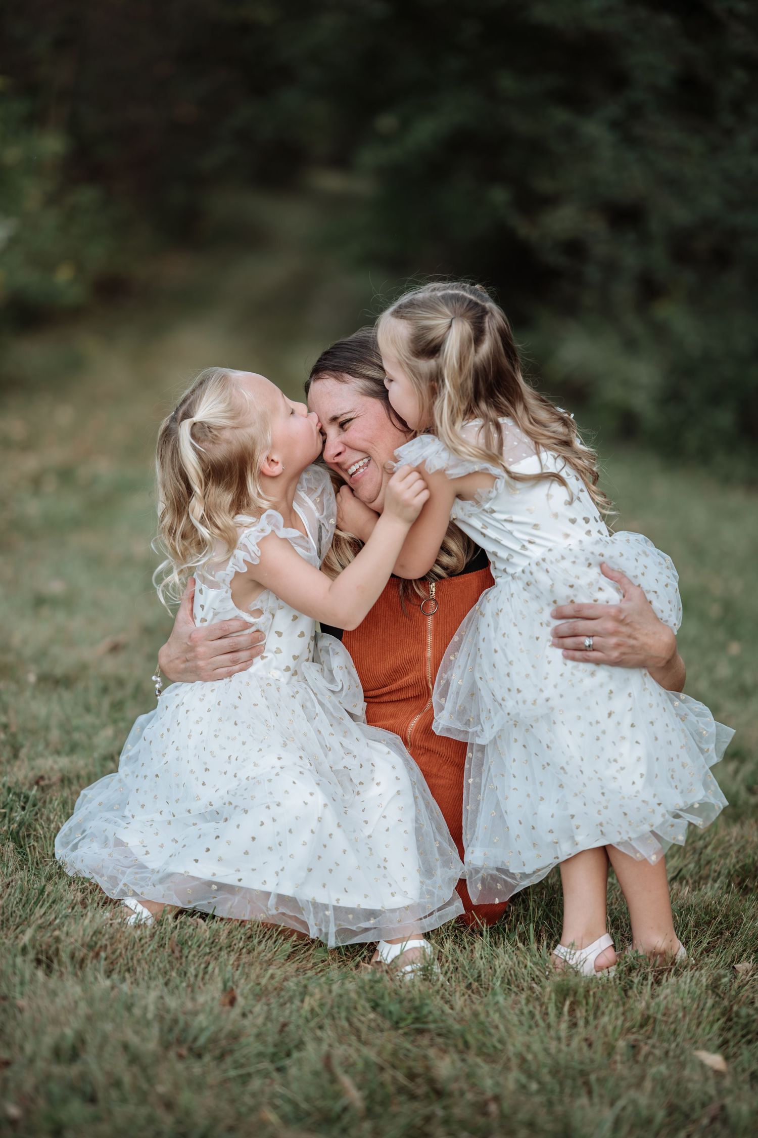 A mother in a rust dress hugs two young girls in white dresses outdoors, sharing a joyful moment together.