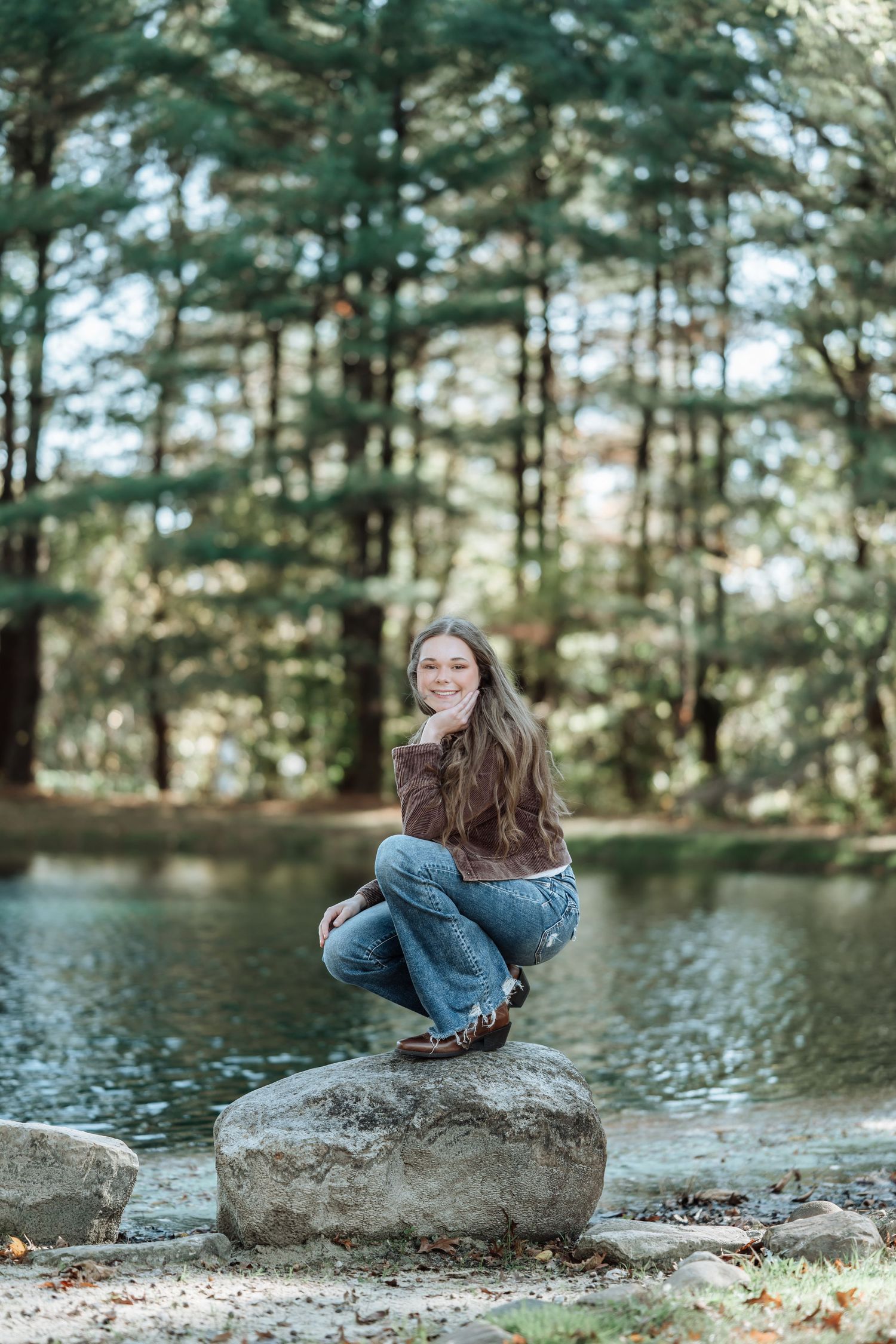 Young woman smiling and posing on a rock by a serene pond surrounded by tall green trees in a forest.