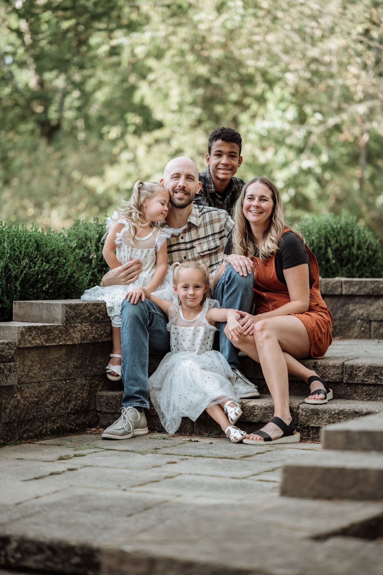 Happy family of five posing on stone steps outdoors, surrounded by lush green trees in a park setting.