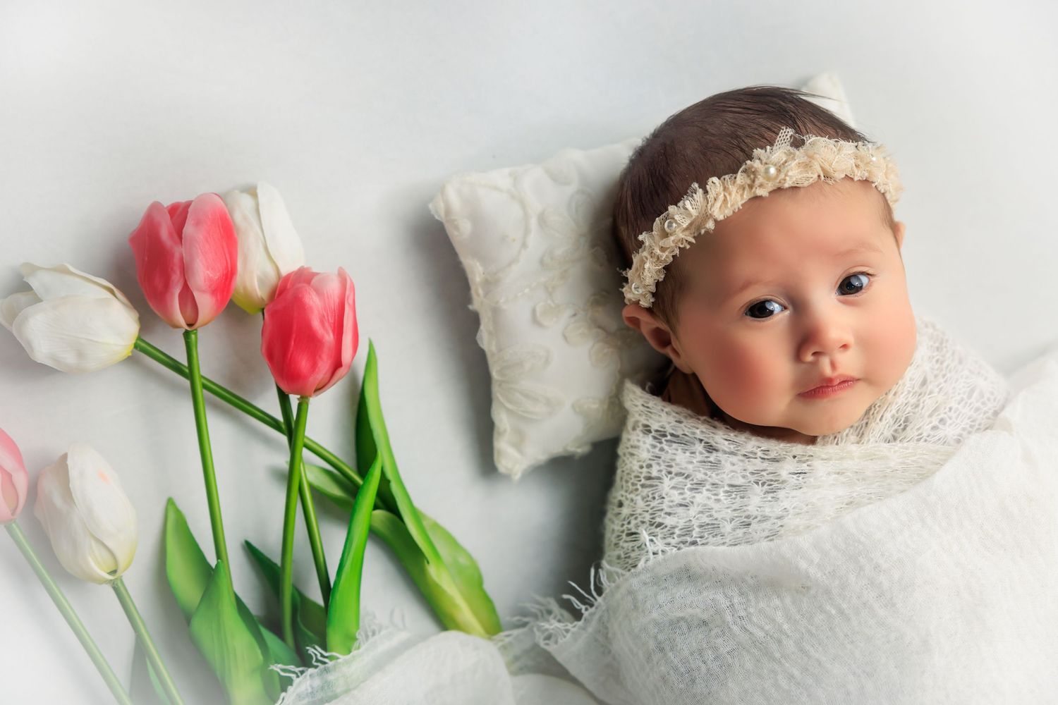 Newborn baby lying on white lace fabric with pink tulips arranged beside them in a professional studio setting.