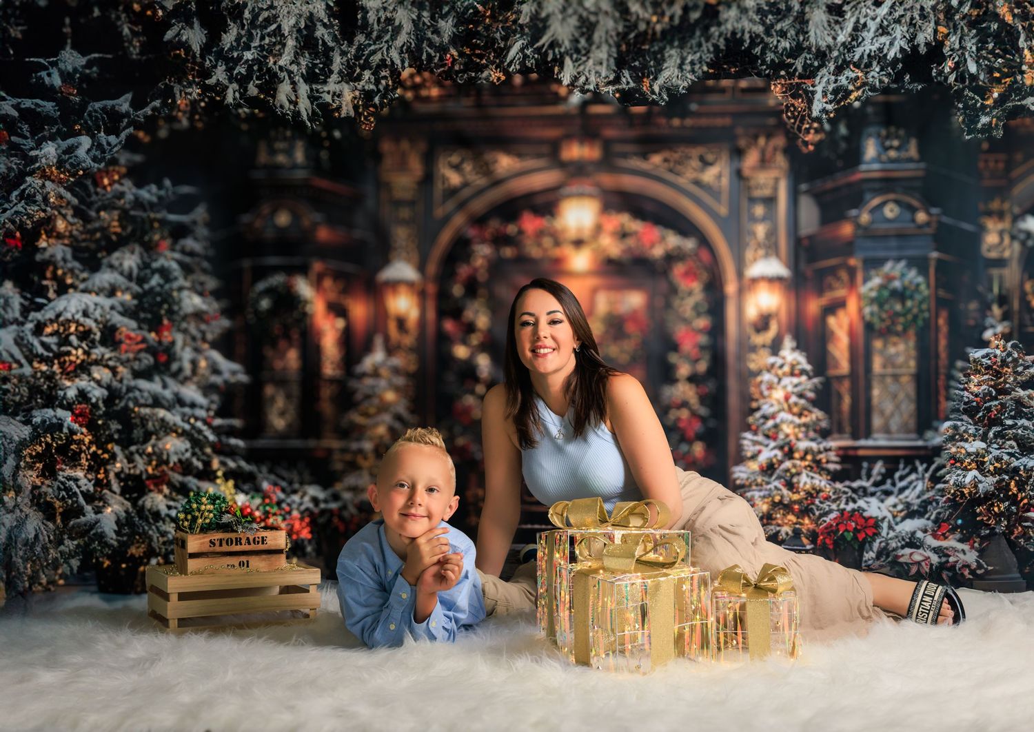 A woman and young boy pose among Christmas trees, gifts, and snow in a festive holiday portrait setting.
