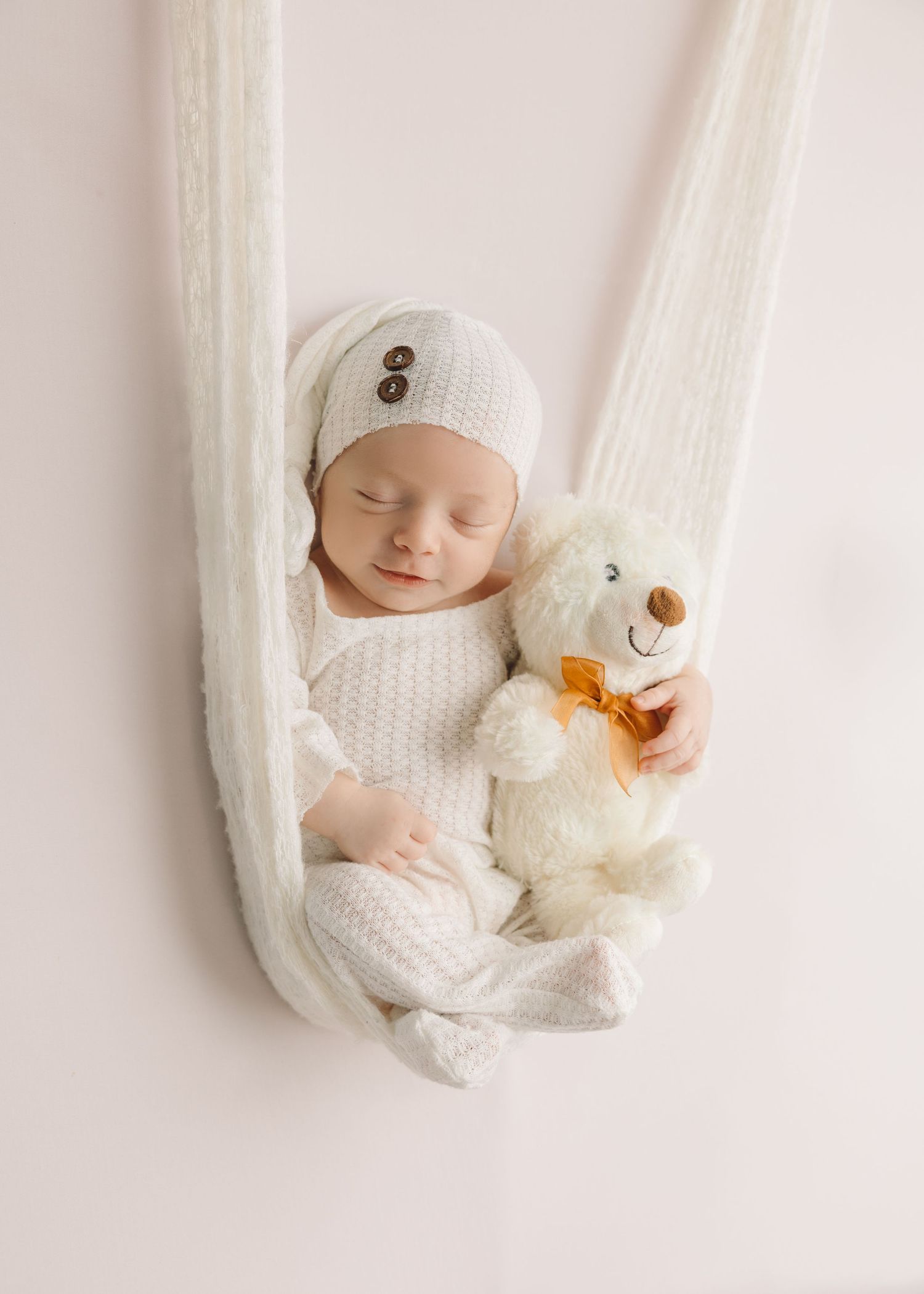 A sleeping baby in white knit clothing lies peacefully in a hammock with a plush teddy bear companion.