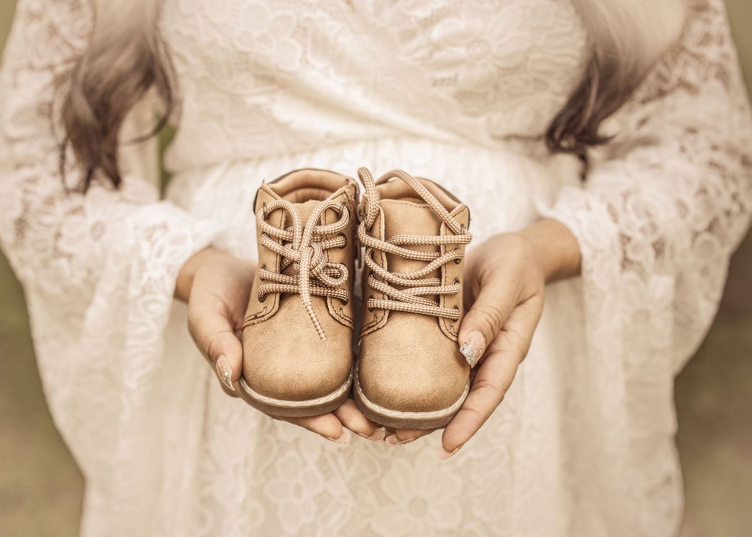 A pair of vintage-style beige baby shoes rests on delicate white lace fabric in a soft, dreamy setting.