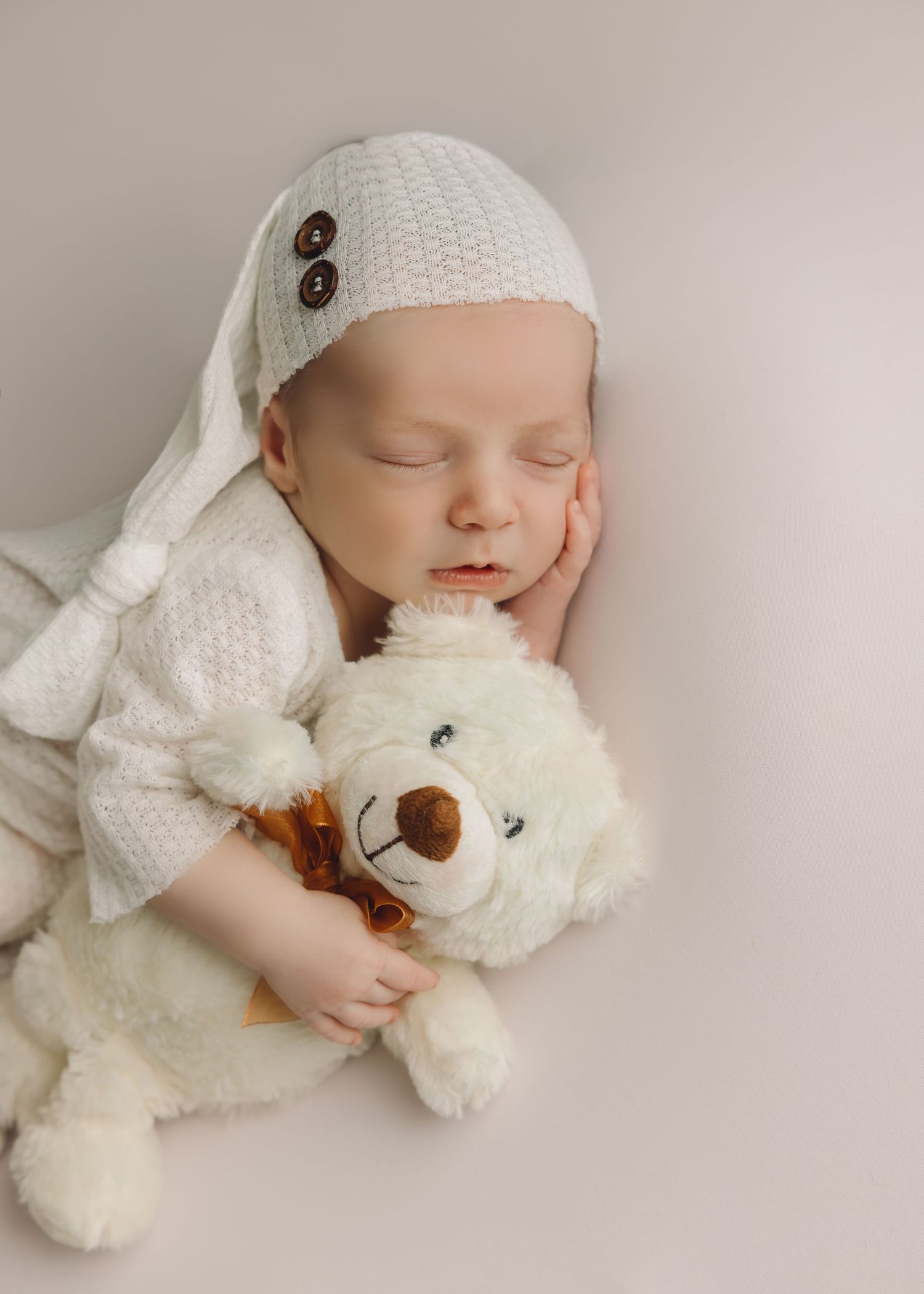 Sleeping newborn in white knit outfit cuddling with soft white teddy bear in gentle studio lighting.