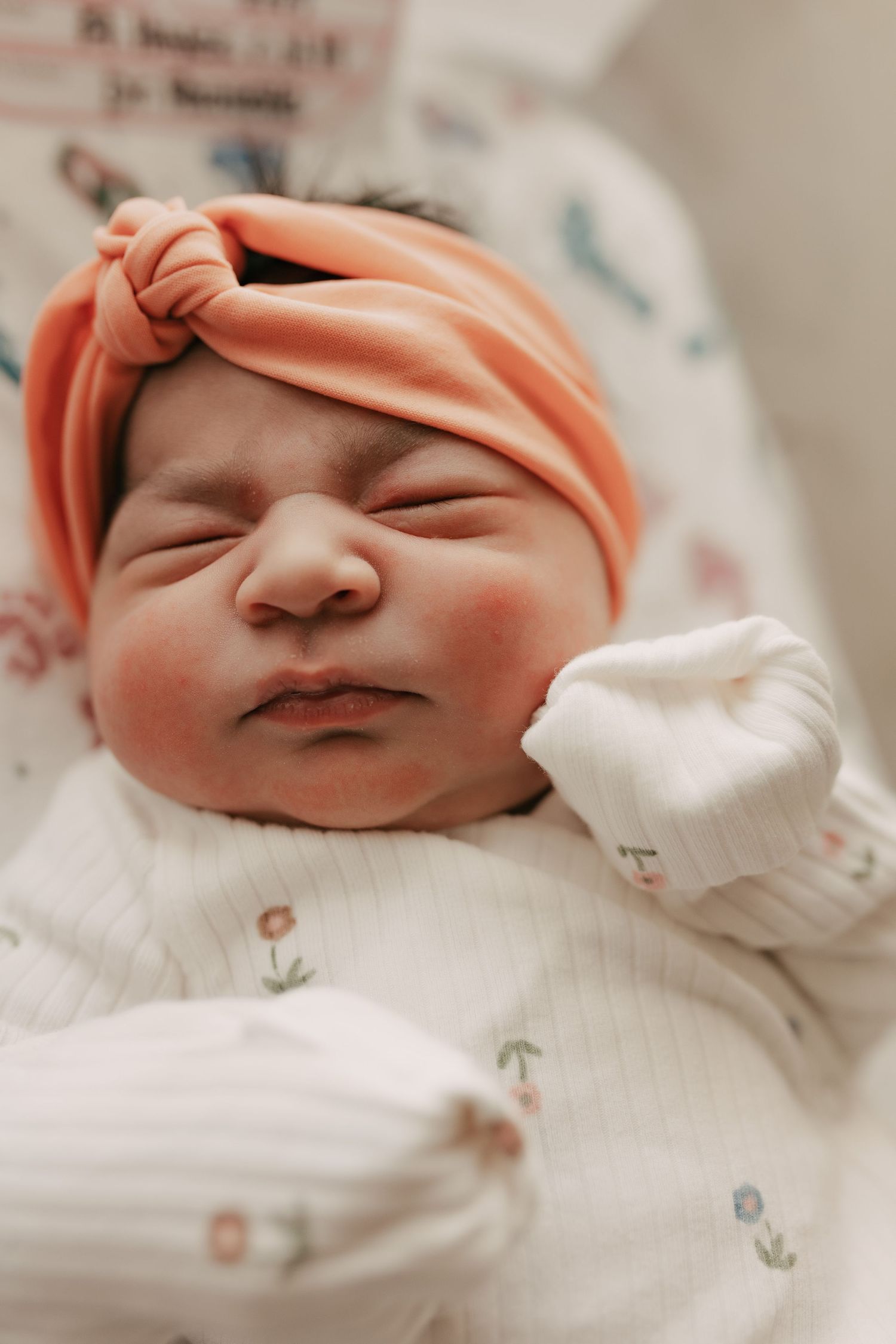 A newborn baby wearing a peach-colored headband and floral onesie sleeps peacefully.