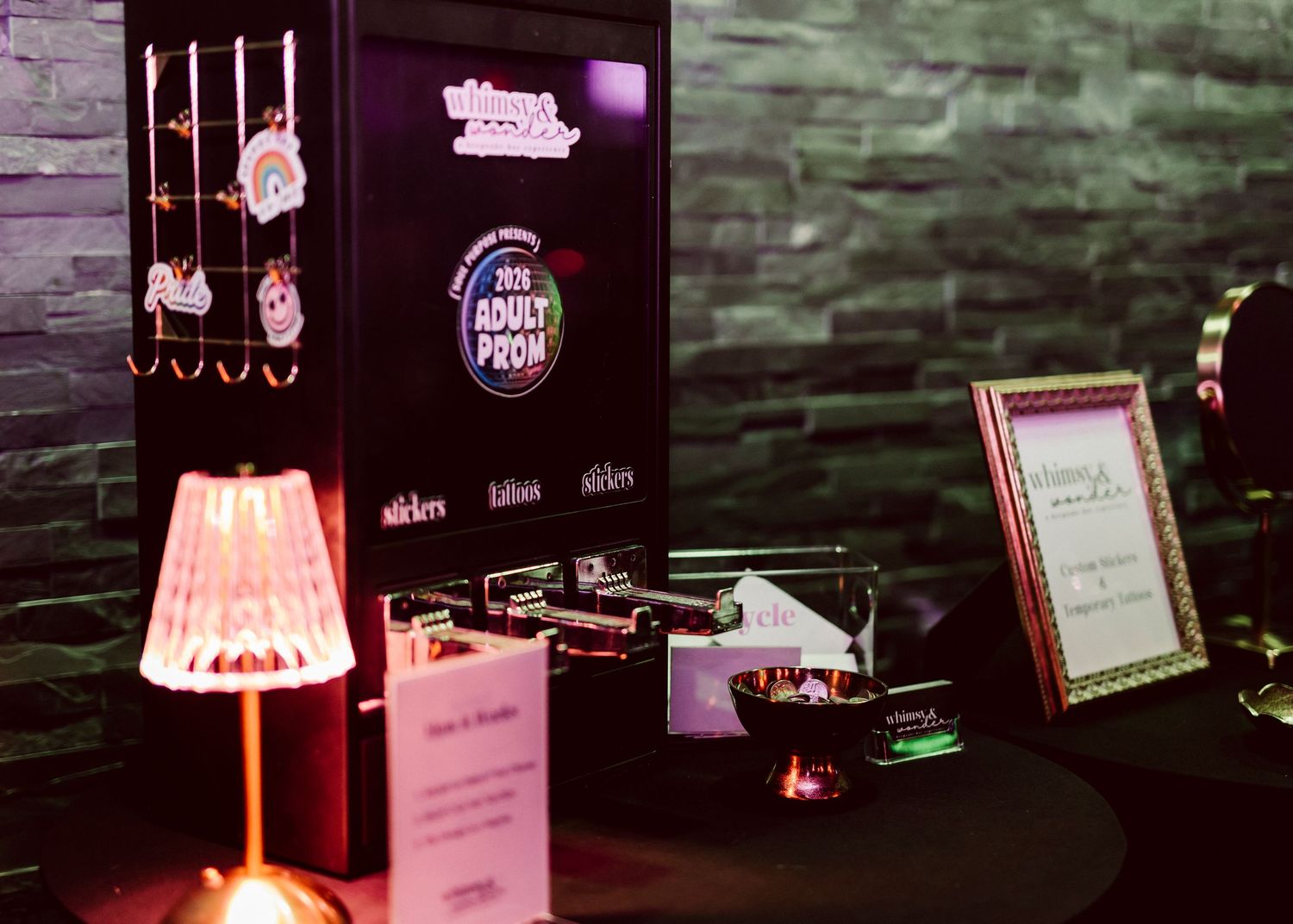 Dark event display booth featuring Adult Prom signage, illuminated lamp, and branded merchandise on a black table.